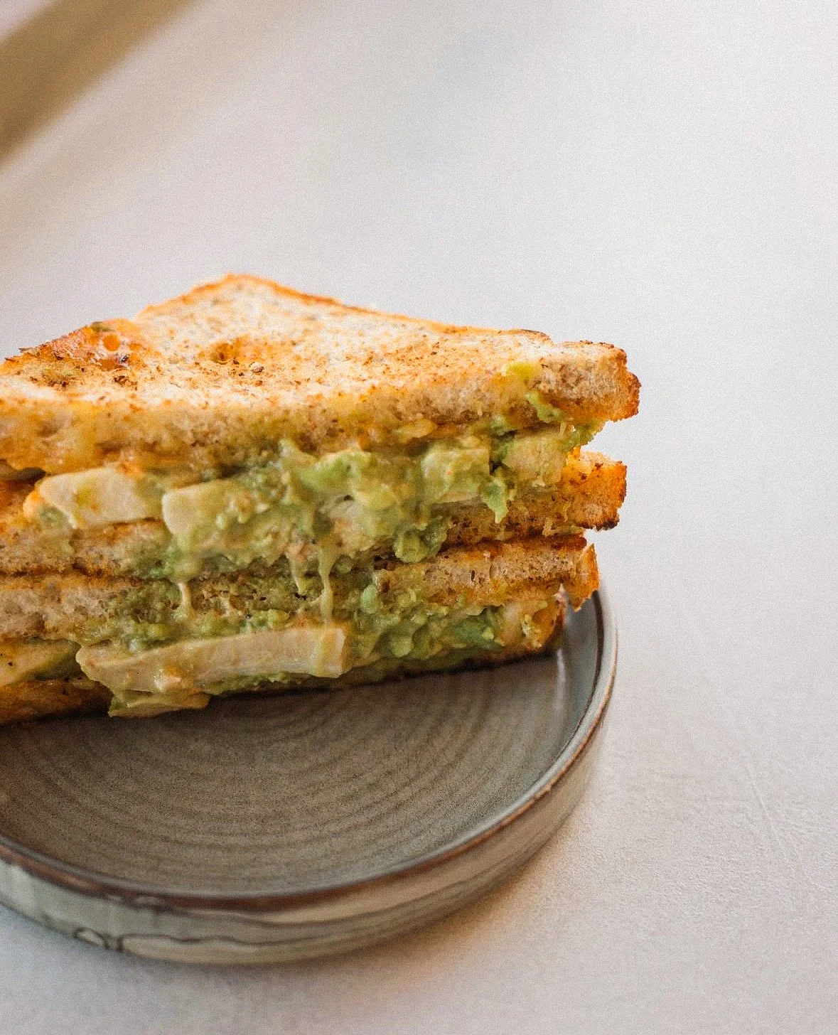 Close-up of a toasted triple-layer sandwich with avocado, onion, and other ingredients on a small ceramic plate.