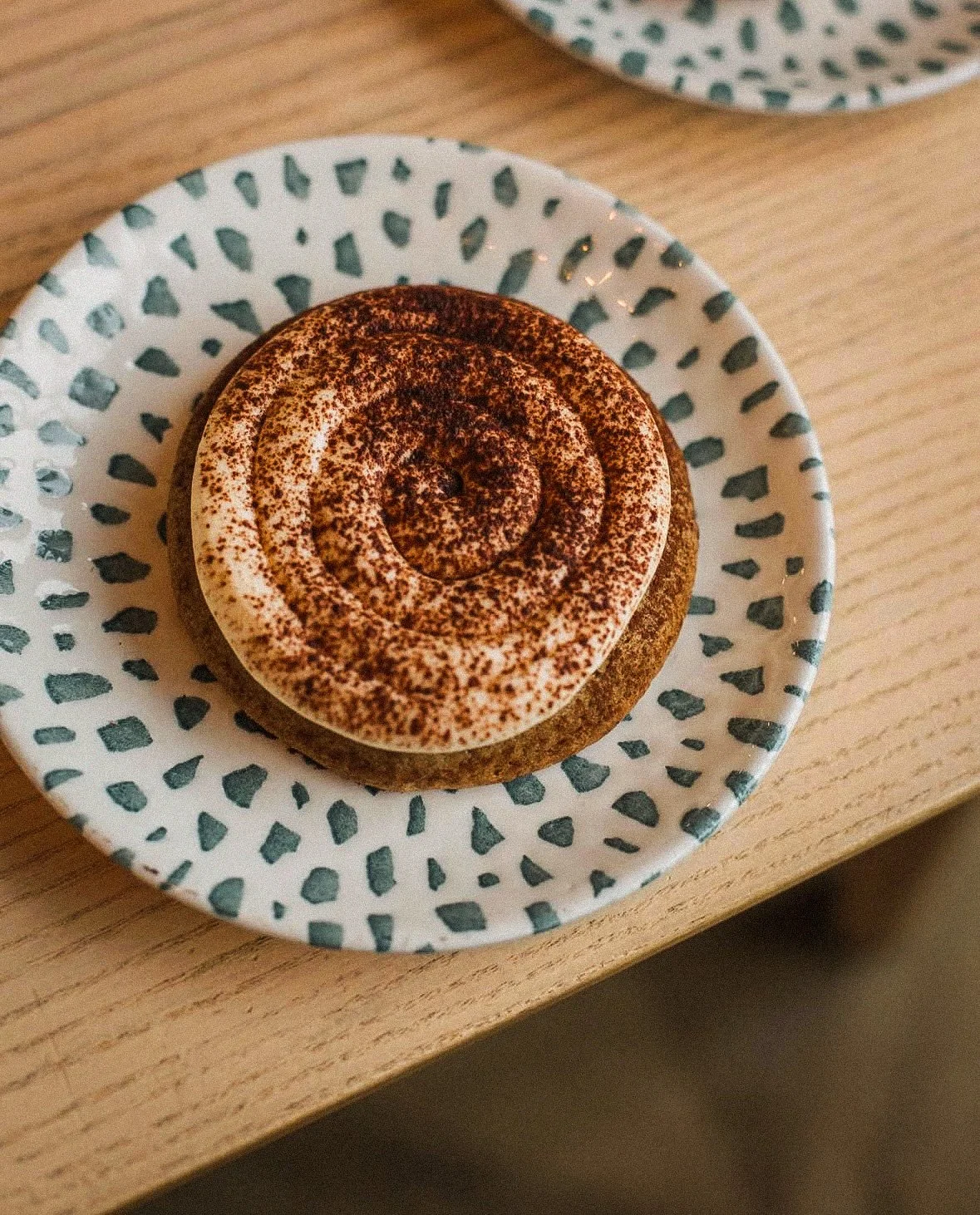 A plate with a cinnamon roll topped with icing and cocoa powder, placed on a wooden table.