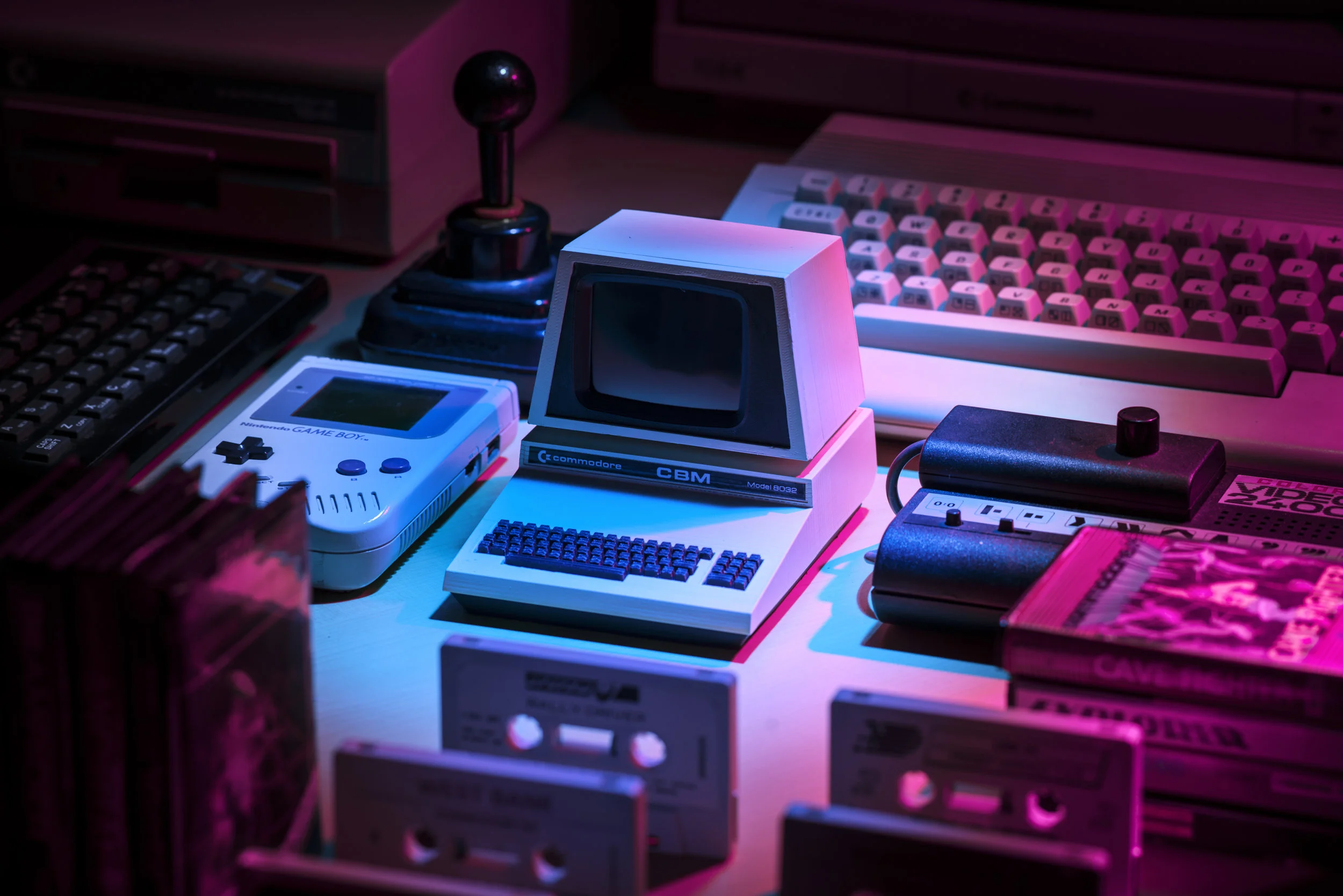 A collection of vintage technology devices on a desk illuminated with pink and blue lighting, including an Nintendo Game Boy, an old computer monitor, a computer keyboard, a joystick, a cassette player, and several cassette tapes.