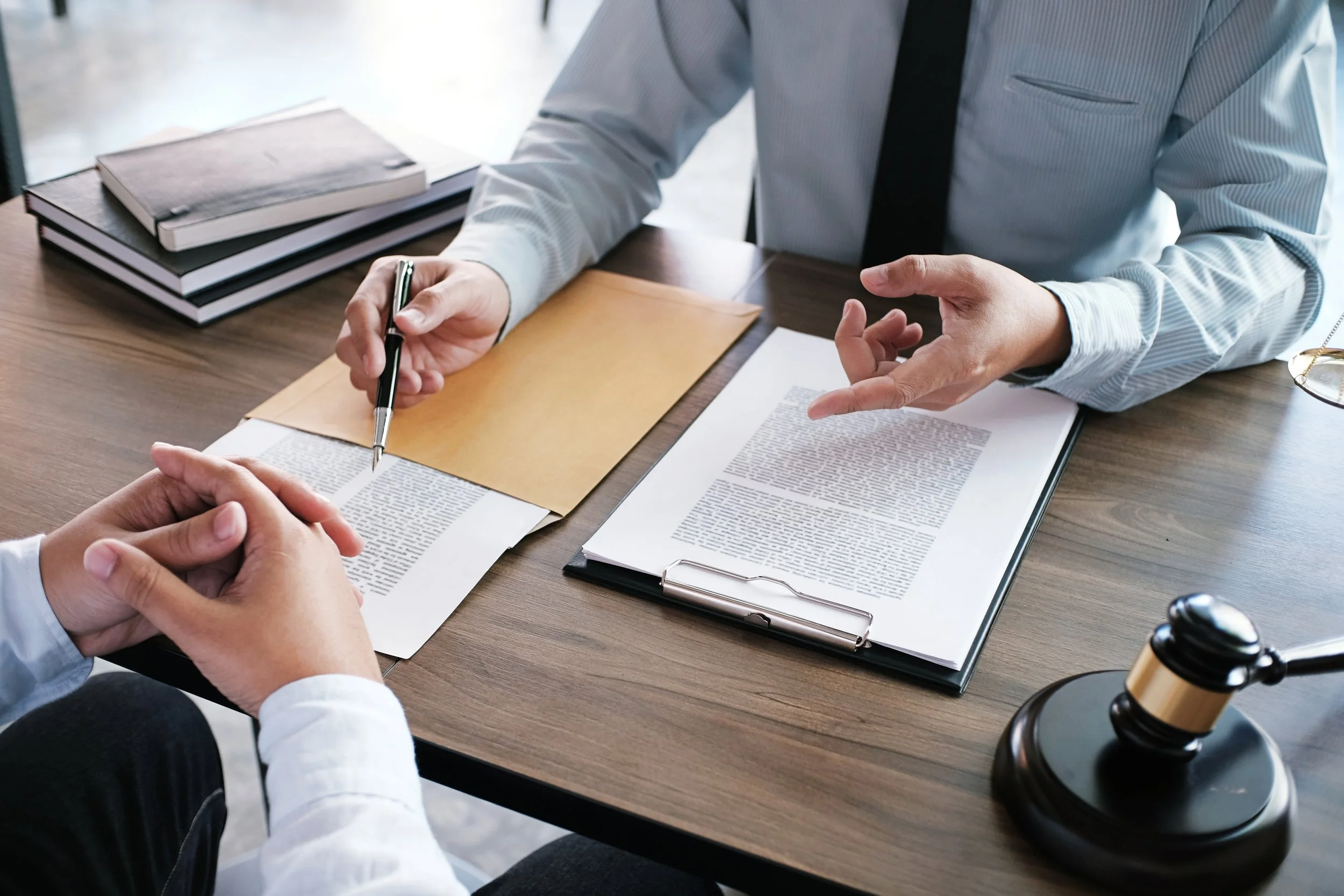 A lawyer or judge in a suit explaining legal matters to a client or colleague during a consultation. The scene includes a gavel, documents, and a folder on a wooden table.