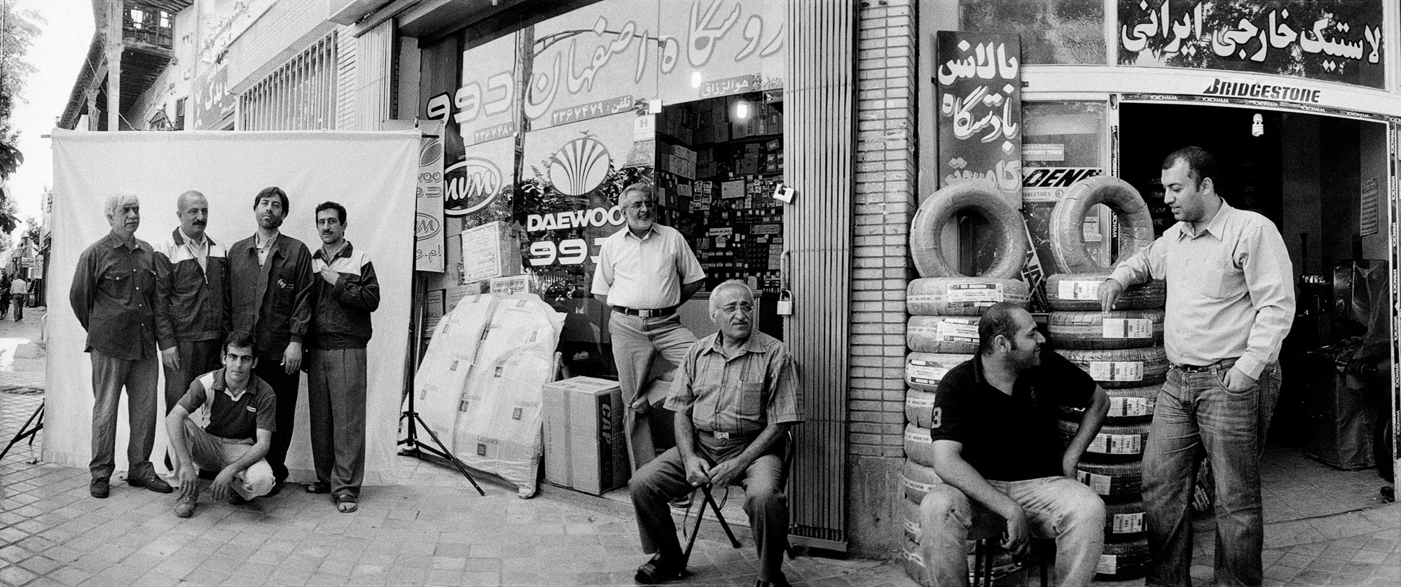 A wide black-and-white panoramic frieze of an Iranian tyre-and-parts street with Persian shop signage and stacked tyres; nine or ten men are strung across the frame — four standing stiffly against a hung white sheet with a fifth crouched at their feet, one standing in a shop mouth, one seated on a stool with a cane, one crouched among stacked tyres, one standing hands-in-pockets at the right edge