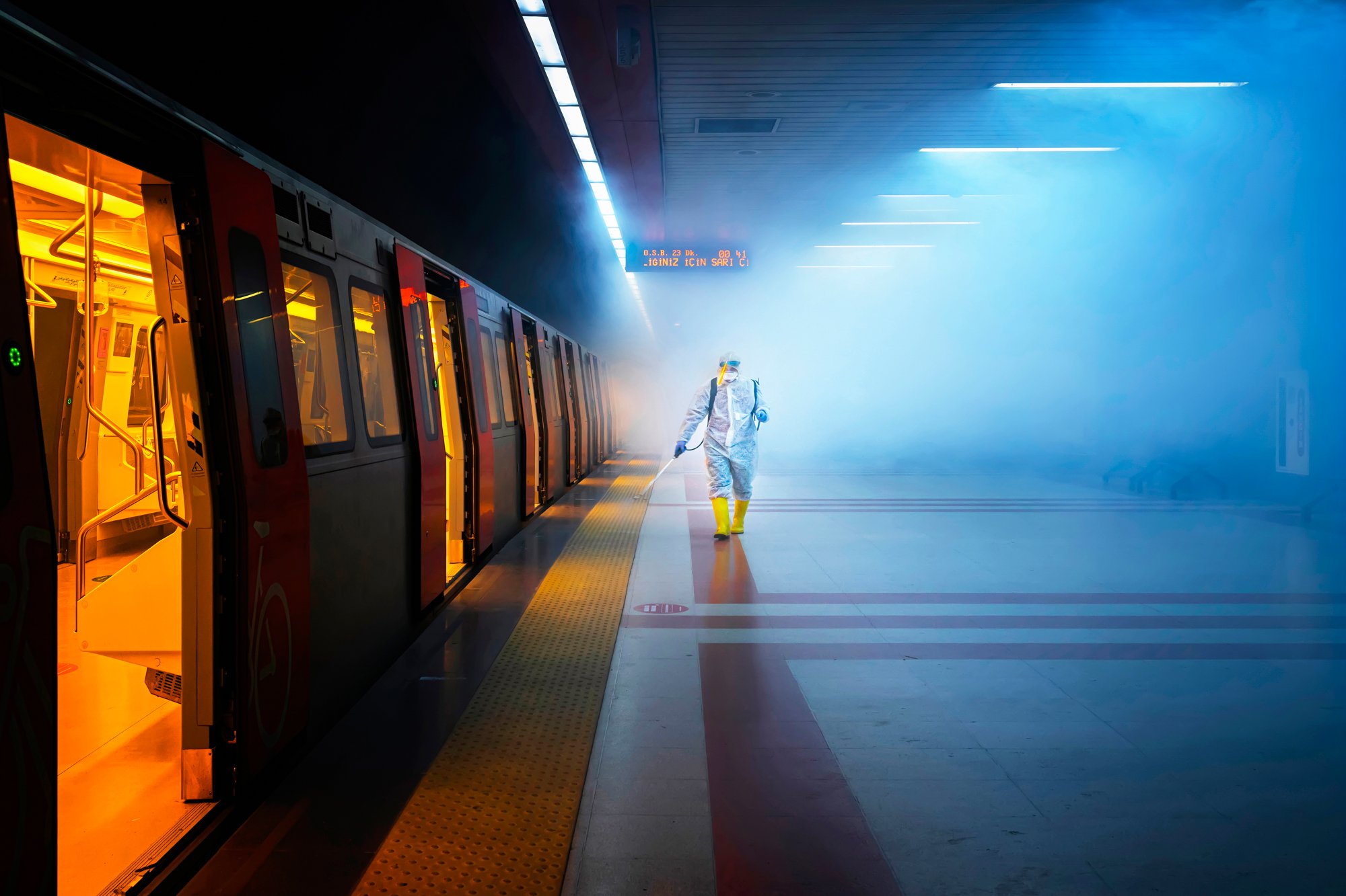 An Ankara metro platform at night: a lone worker in a white hazmat suit and yellow rubber boots walks away spraying disinfectant into a wall of cold blue haze, with an open orange-lit carriage running down the left edge