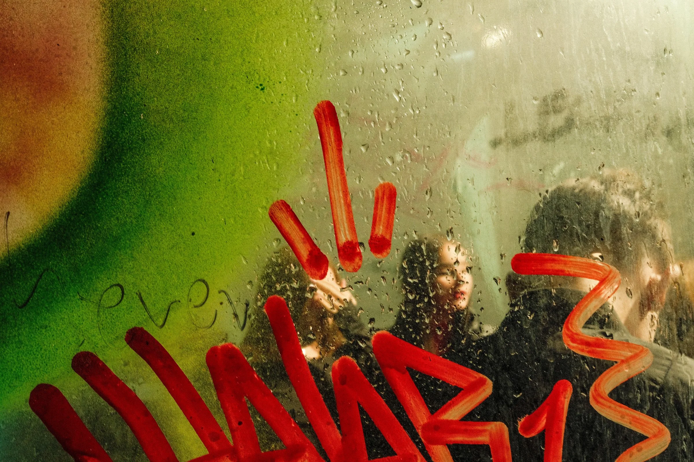 Shot through a steamed, rain-beaded pane scribbled with thick red finger-strokes and the word "even"; behind the glass a green-and-orange wash and three or four blurred passengers, one woman's face readable through a clear patch