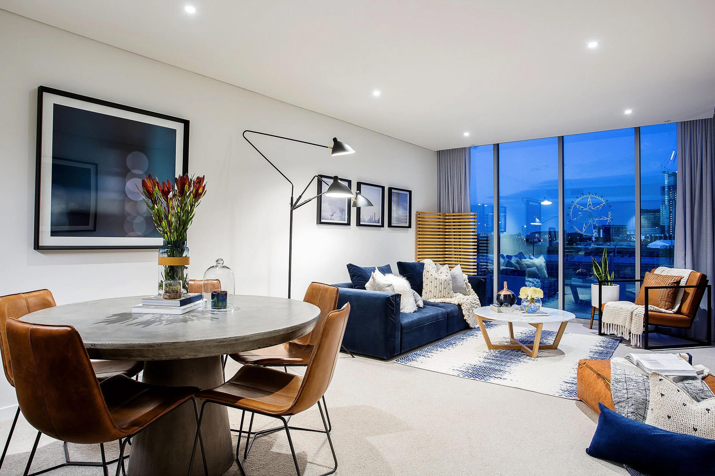 Open plan living and dining area in Forge by Mirvac Apartment 204, Docklands, styled with navy sofa, leather dining chairs and floor to ceiling glazing overlooking Melbourne skyline.