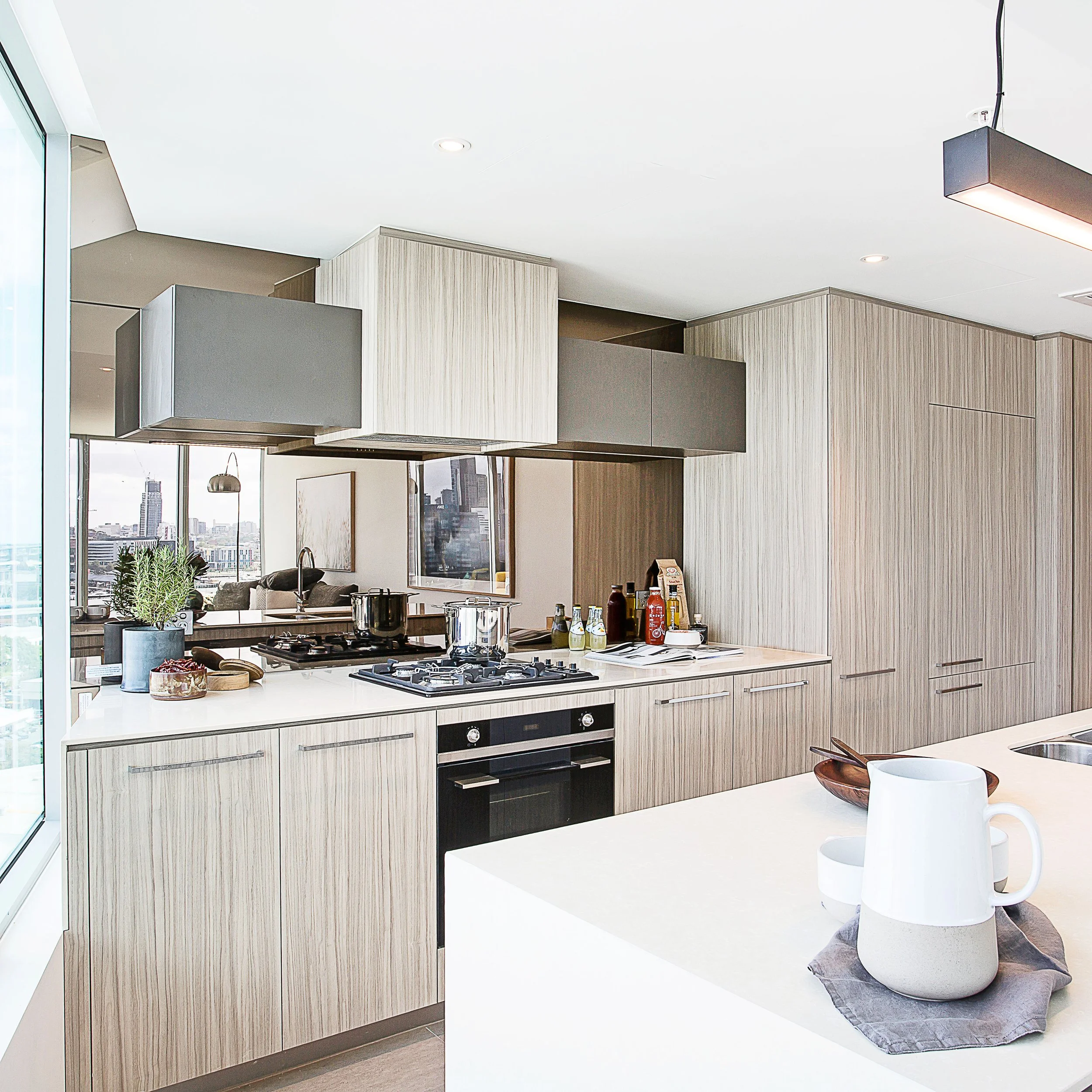 Modern kitchen in Mirvac Forge apartment with timber veneer cabinetry, stone island bench and mirrored splashback reflecting city skyline.
