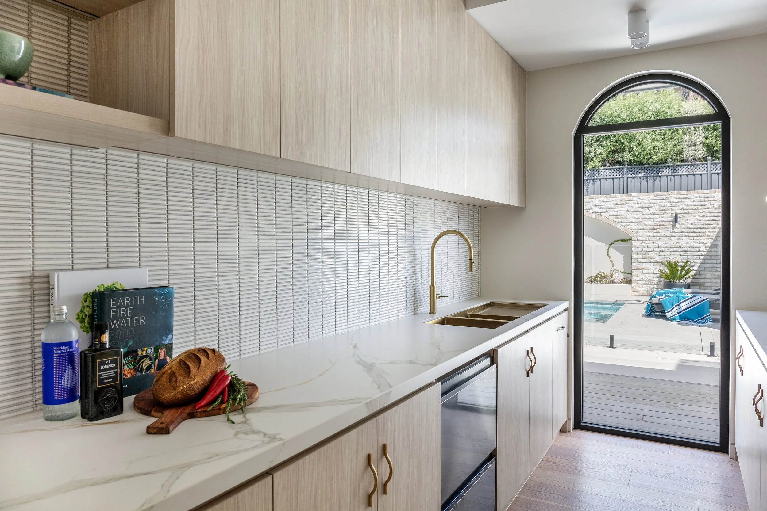 Butler’s pantry at Dromana residence with brass sink, stone benchtop, pale timber cabinetry and view to pool