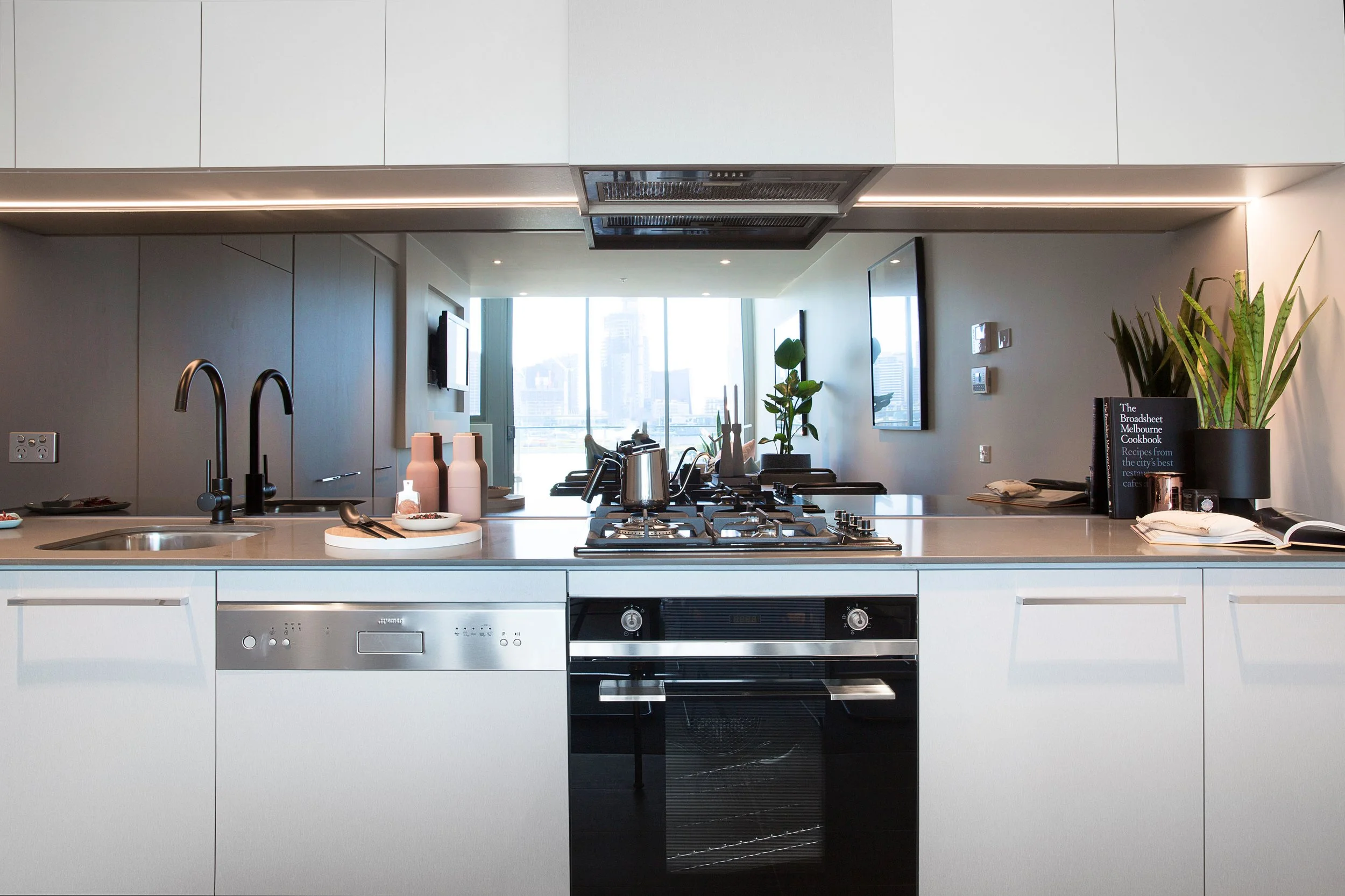 Streamlined white kitchen with mirrored splashback and integrated appliances, styled to emphasise space and light in a contemporary Melbourne loft apartment.