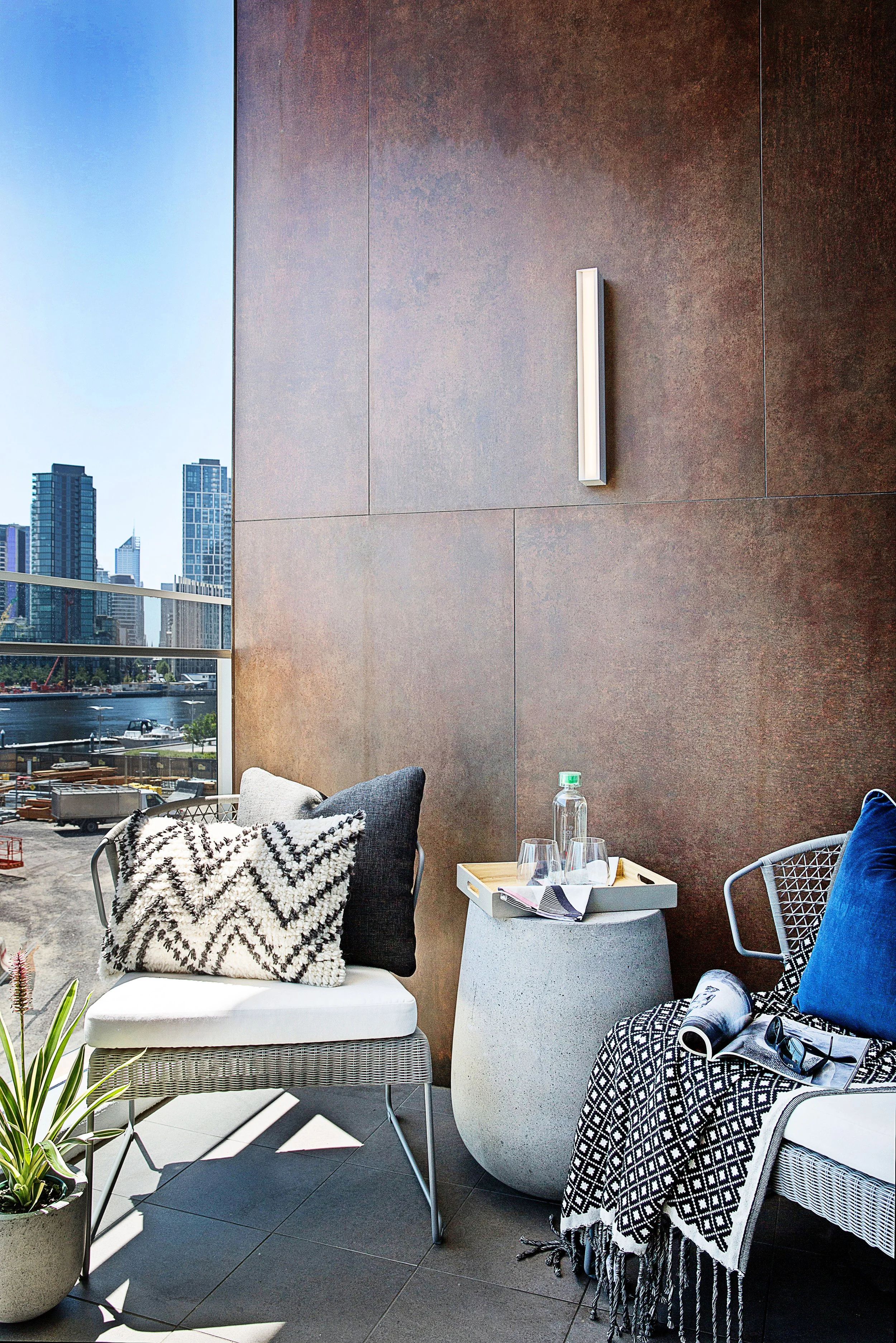 Outdoor terrace styled as an urban retreat with textured cushions, outdoor armchairs and city skyline views at Forge, Yarra’s Edge. A sophisticated loft-style apartment interior by DMP Creative.