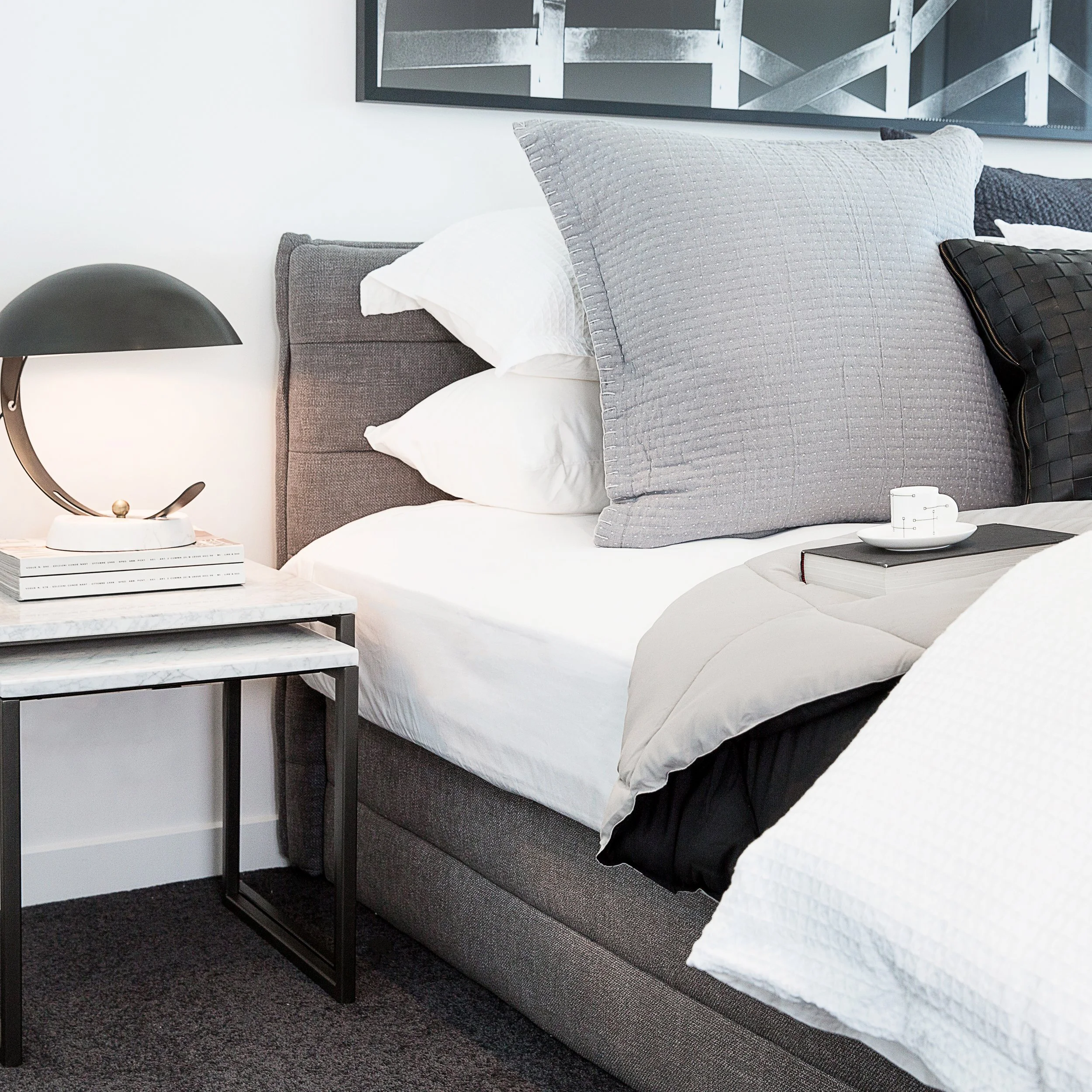 Contemporary loft bedroom styling at Forge featuring layered grey bedding, upholstered bedhead and sculptural bedside lamp. Designed to evoke relaxed urban luxury for sophisticated Melbourne apartment buyers.