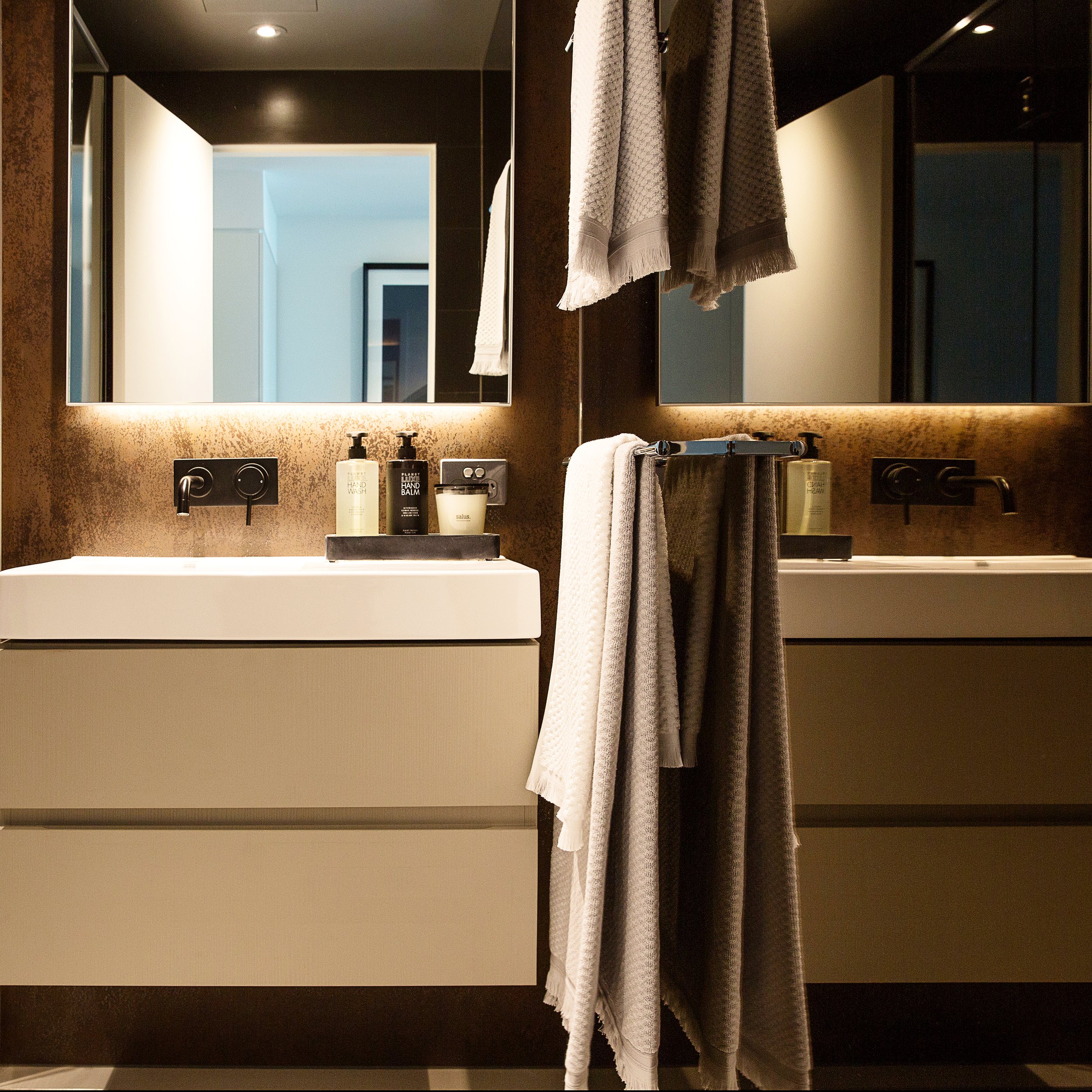 Custom styled loft bathroom at Forge Yarra’s Edge featuring double vanity, warm bronze mirror wall, under-cabinet lighting and layered neutral textures. A refined, hotel-inspired ensuite designed for sophisticated inner-city living.