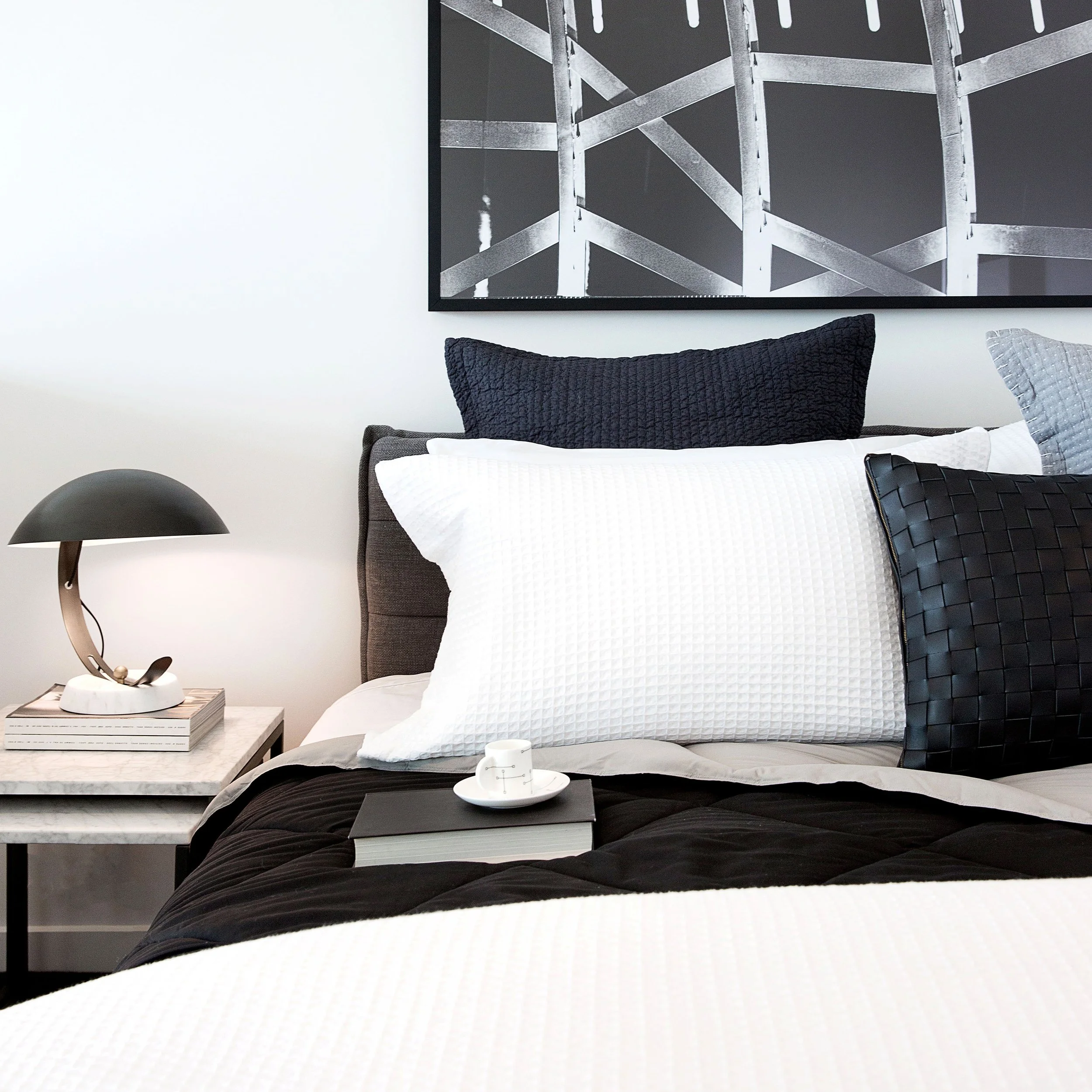 Loft apartment bedroom styled in a monochrome palette with charcoal upholstered bedhead, layered white and black bedding, marble bedside table and contemporary Melbourne artwork above bed.