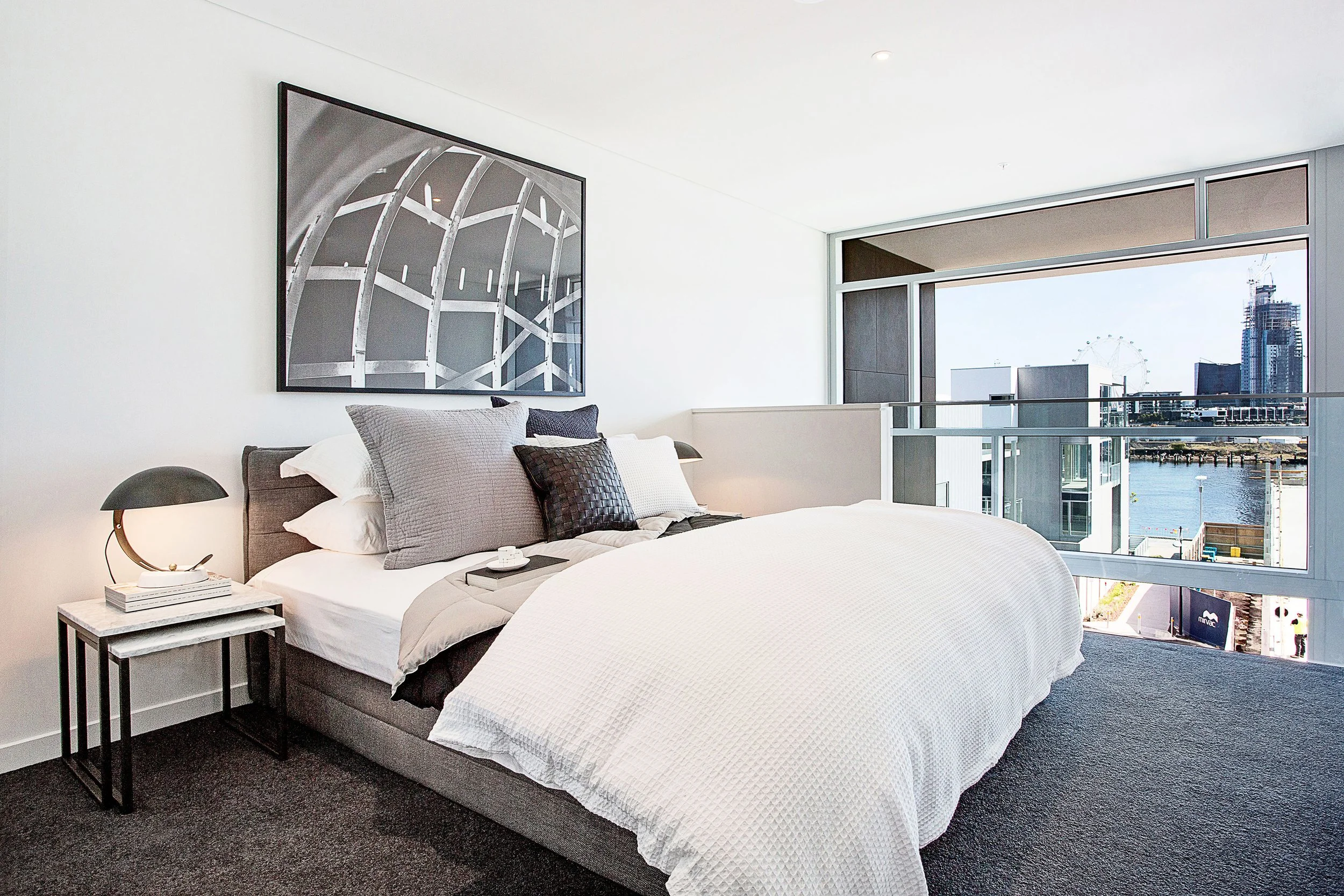 Elevated loft bedroom overlooking Melbourne skyline, styled in soft neutrals and layered textiles to balance industrial architecture with comfort.