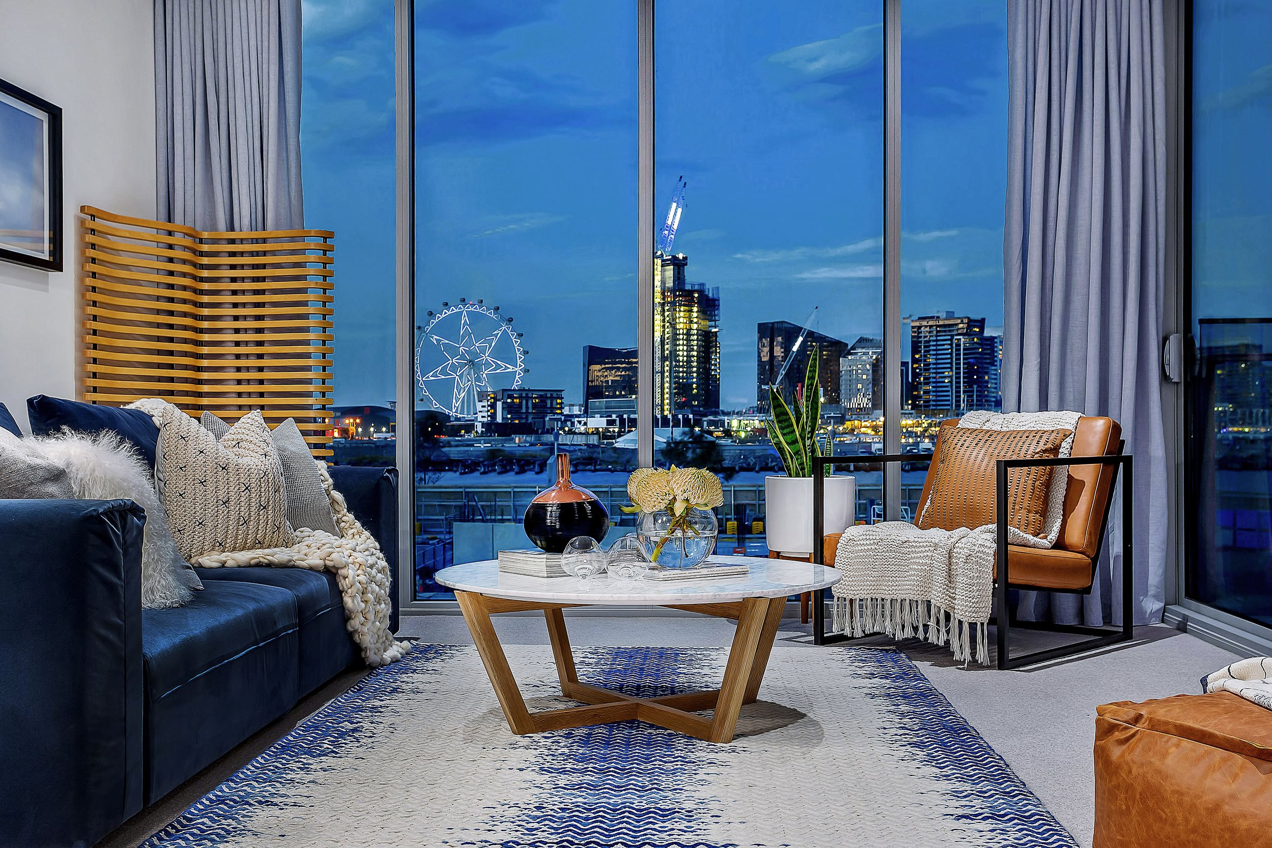 Living room in Forge Docklands Apartment 204 at night, styled with layered textiles and marble coffee table, framed by city skyline and Melbourne Star views through full height windows.