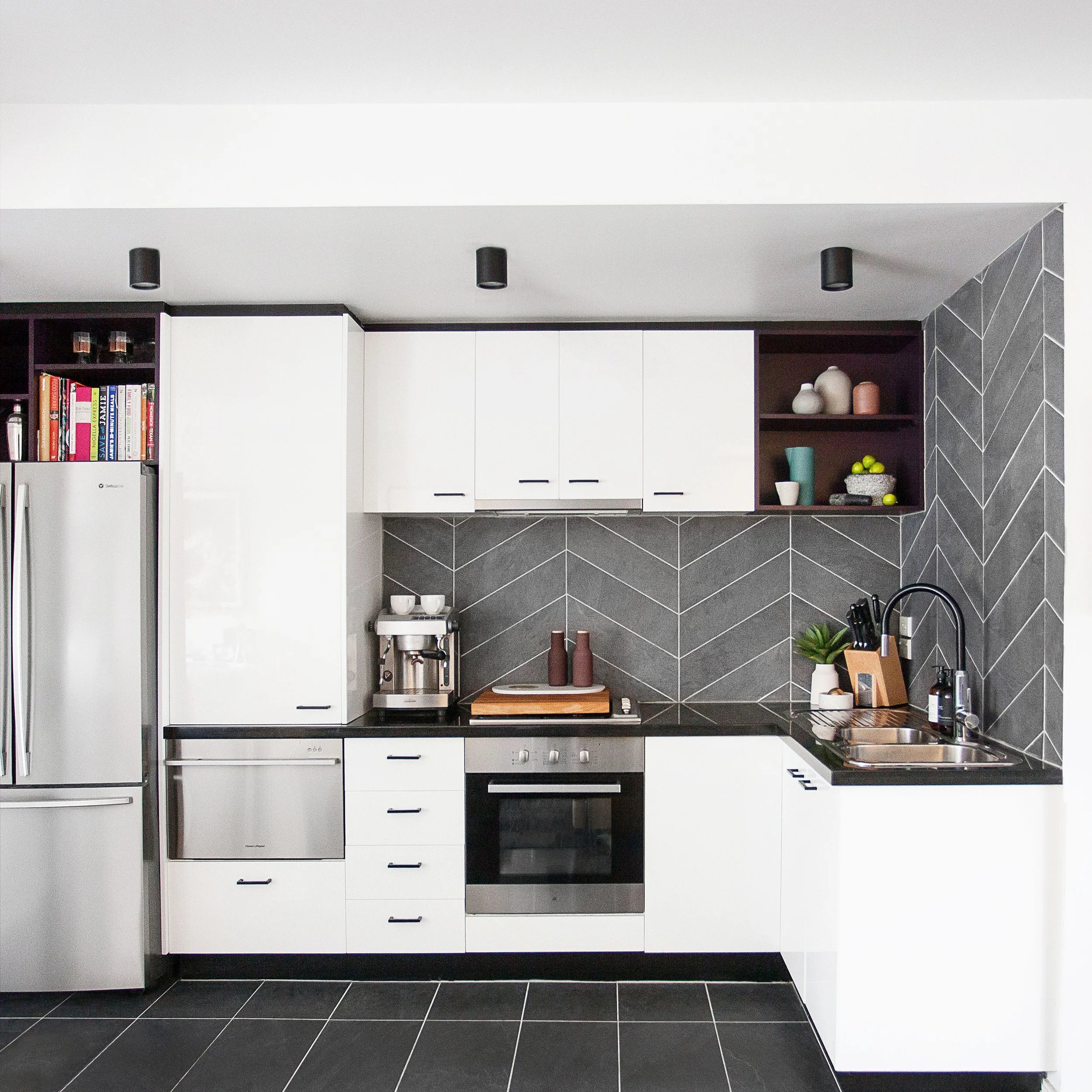 Compact apartment kitchen renovation featuring white joinery, chevron tiled splashback and dark benchtops in a Melbourne inner-city apartment.