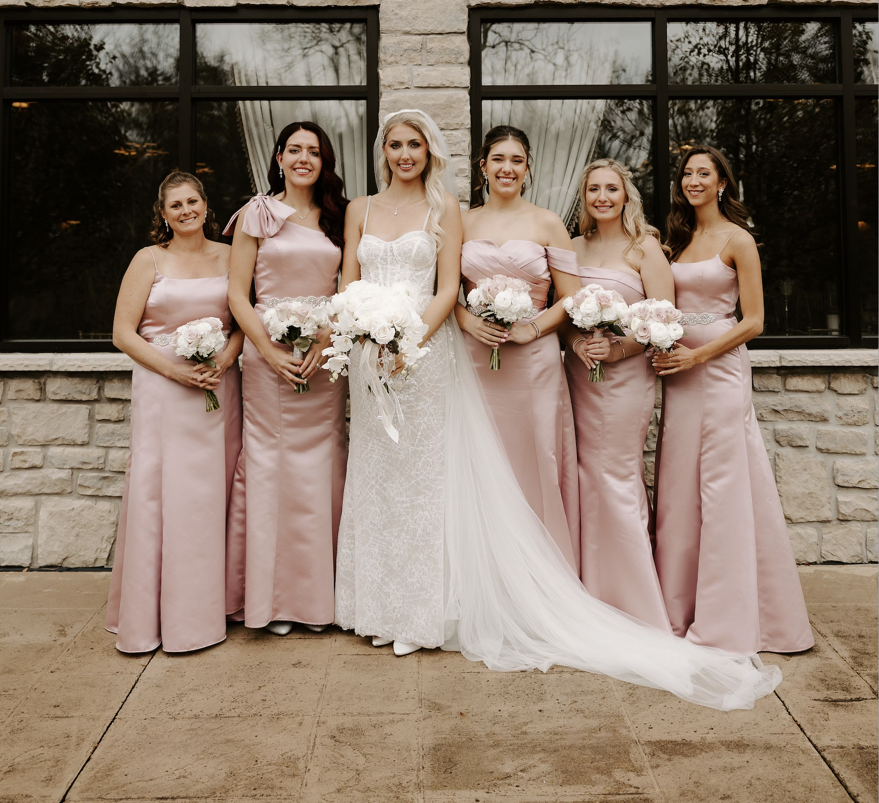 Group of bridesmaids and a bride standing outside in front of a brick building with windows, holding bouquets of flowers, dressed in pink gowns with the bride in a white wedding dress with a train.