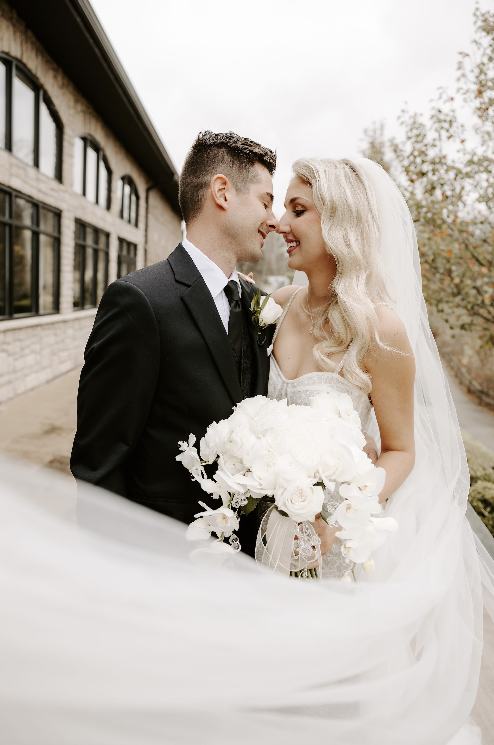 A bride and groom on their wedding day, smiling and leaning noses together outdoors, with the bride holding a bouquet of white flowers.