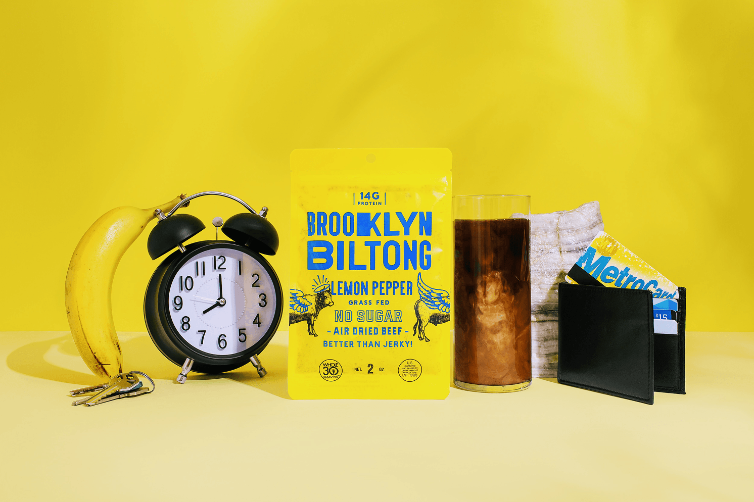 Bright yellow Brooklyn Biltong Lemon Pepper packaging with blue typography, styled with a banana, alarm clock, iced coffee, and wallet against a yellow background.