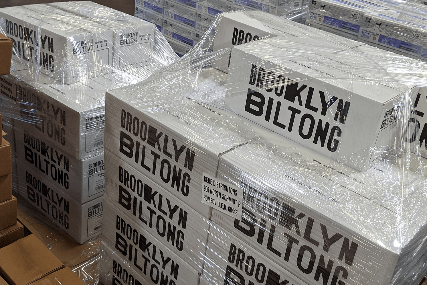 Stacks of Brooklyn Biltong branded shipping boxes wrapped on warehouse pallets, featuring oversized black typography on white cardboard.