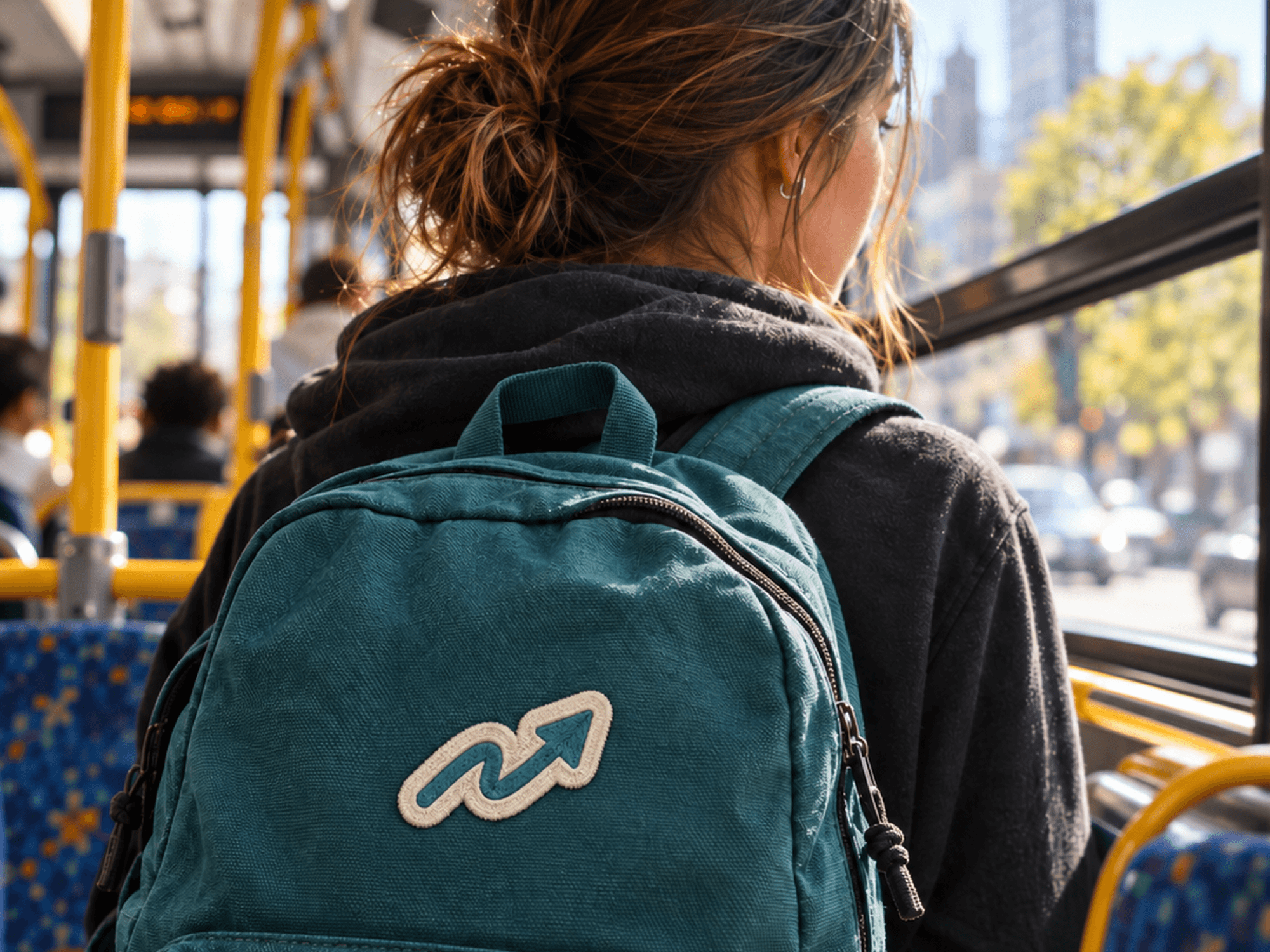 Embroidered backpack: Teal backpack featuring an embroidered Renew arrow logo with a cream outline, worn by a student riding public transportation.