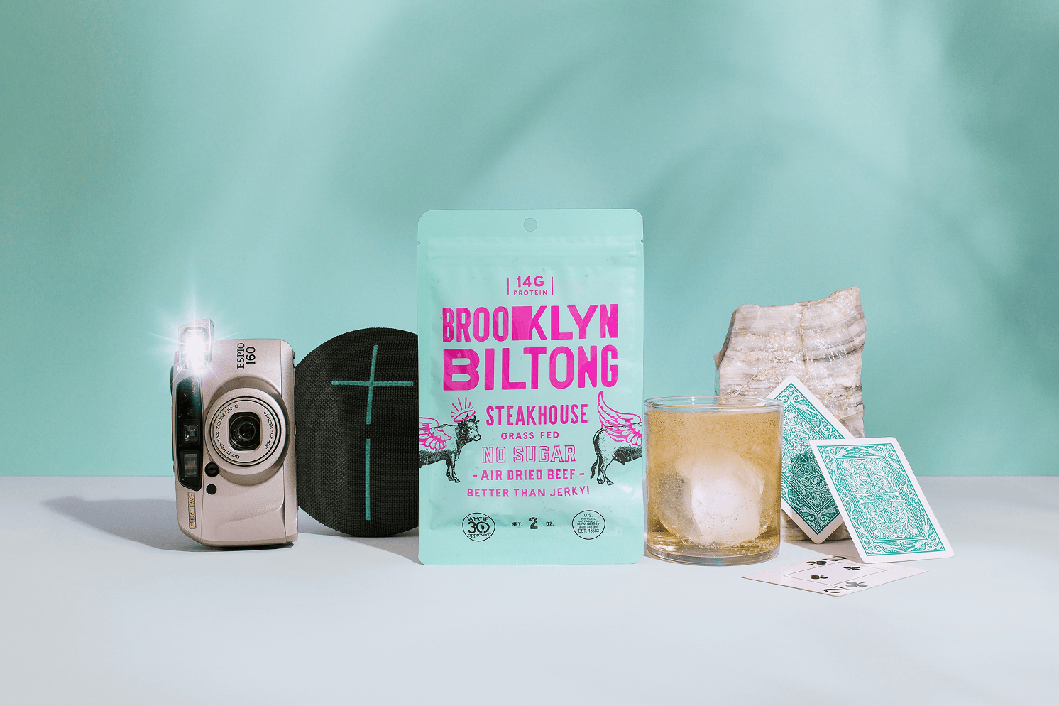 Mint green Brooklyn Biltong Steakhouse packaging with bright pink typography, styled with a camera, speaker, drink, and playing cards.