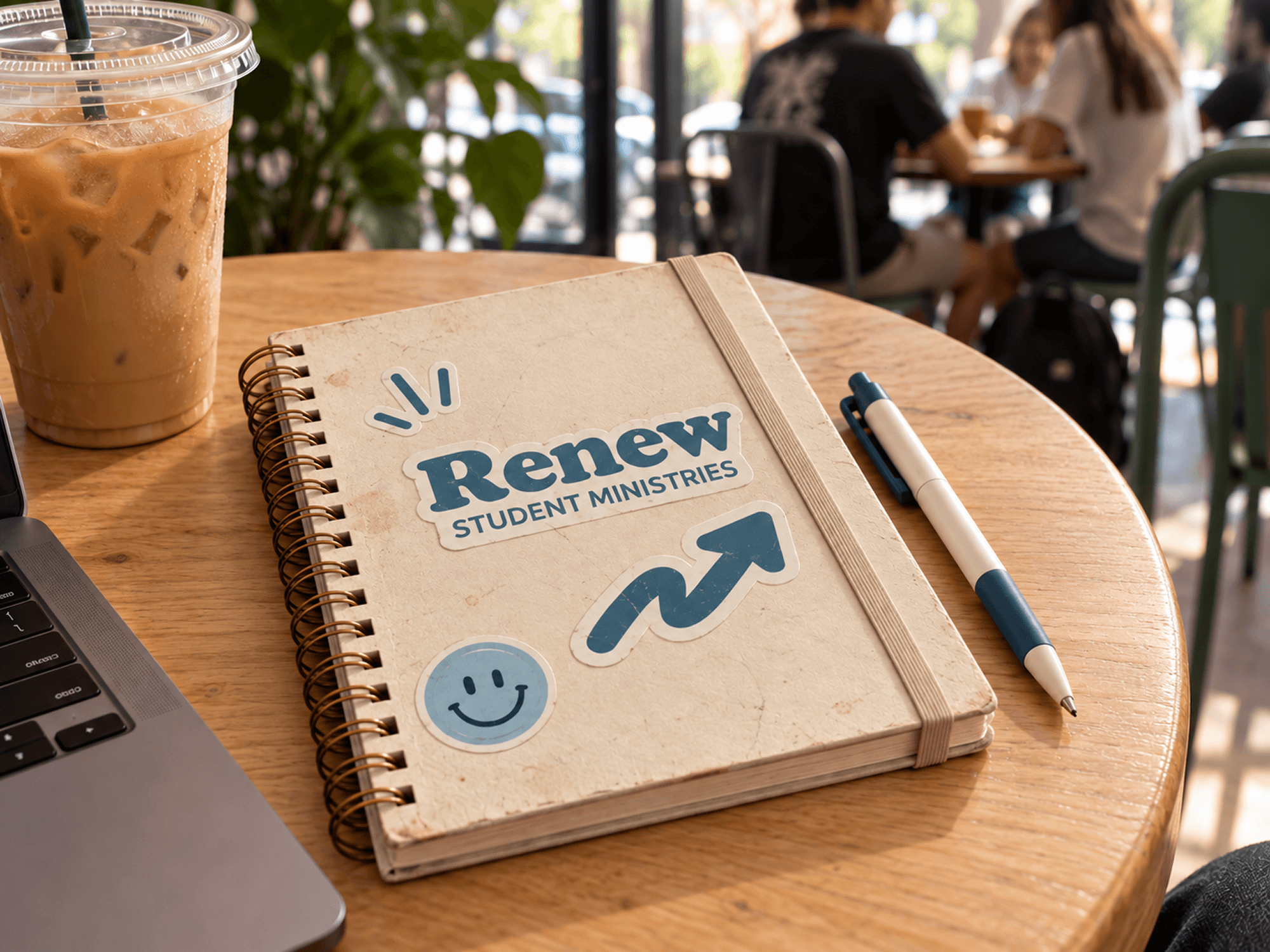 Notebook stickers: Renew Student Ministries logo and arrow stickers displayed on a spiral notebook beside an iced coffee and pen.