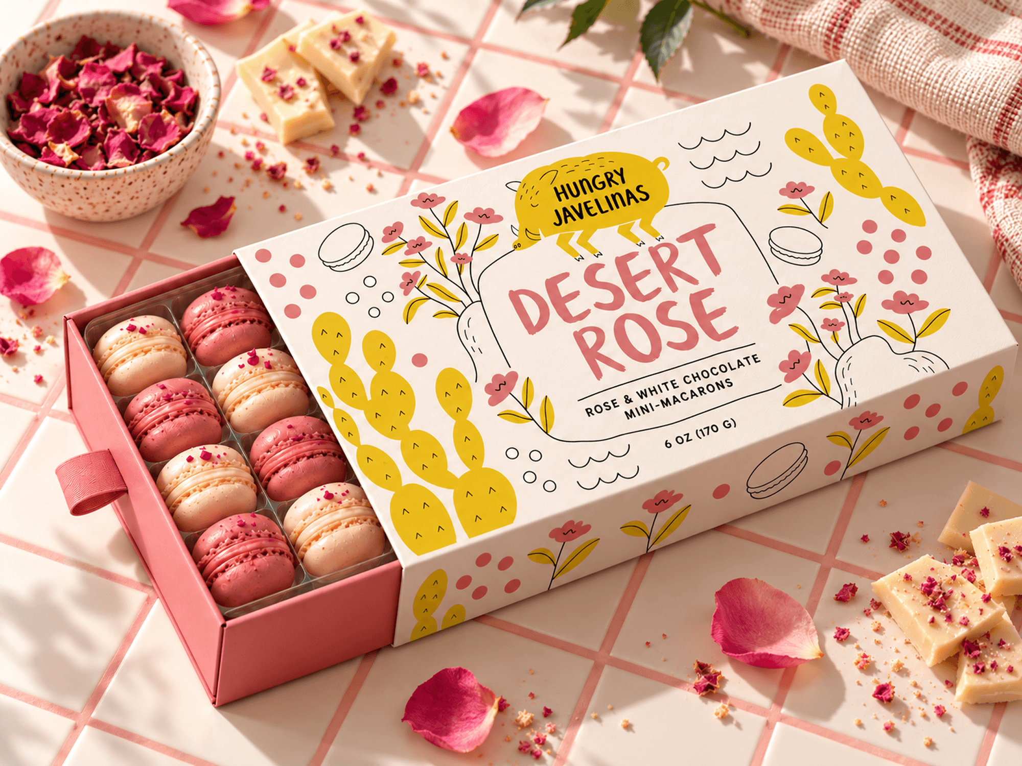 Open Hungry Javelinas Desert Rose mini-macaron box displaying pink and cream macarons, surrounded by rose petals and white chocolate on a pink-grouted tiled countertop.