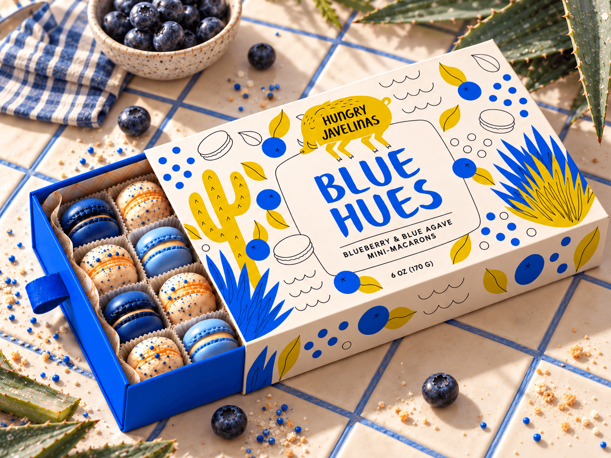 Open Hungry Javelinas Blue Hues box displaying blue and cream mini-macarons, surrounded by blueberries and agave leaves on a blue-grouted tiled countertop.