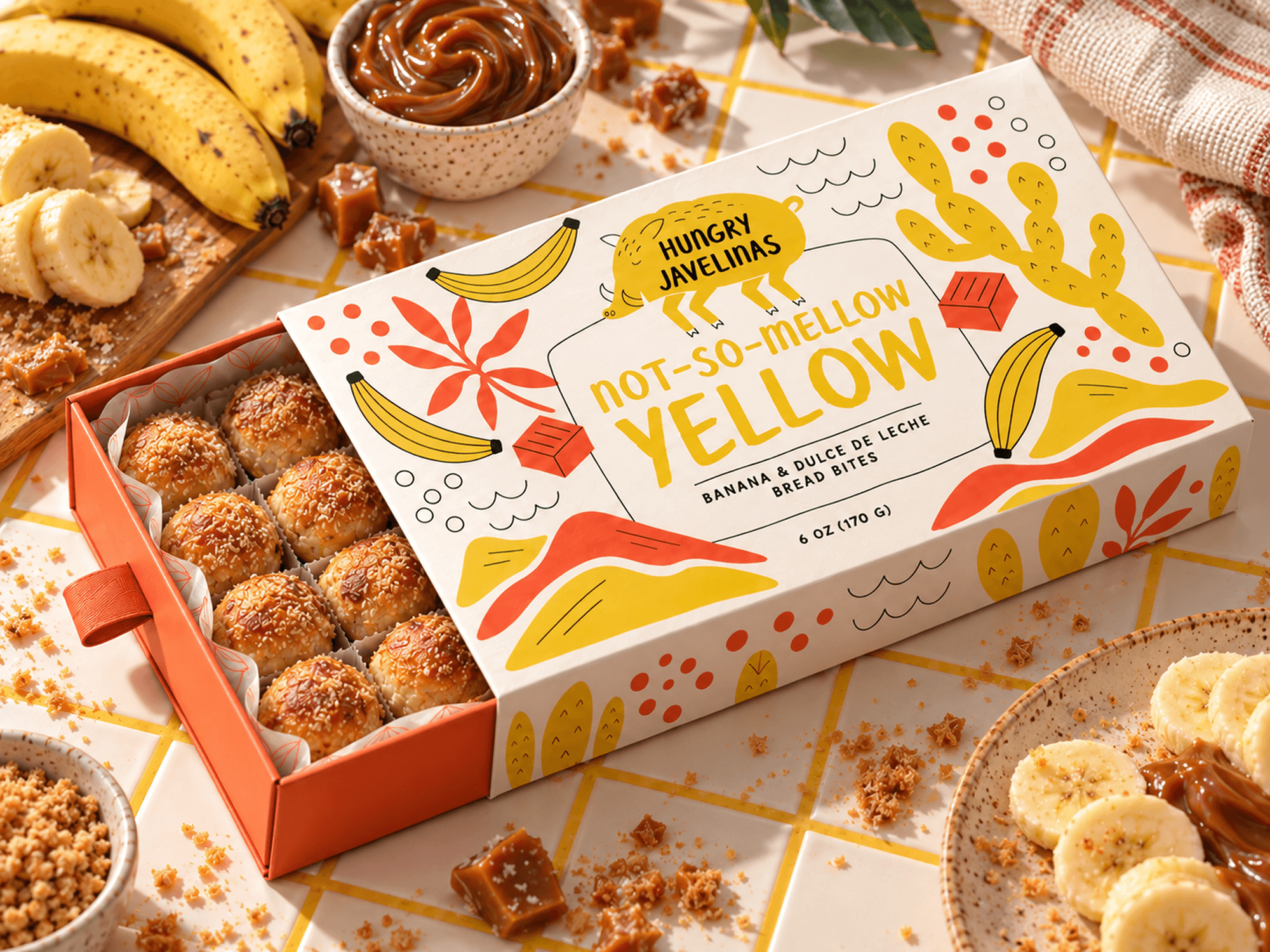 Open Hungry Javelinas banana and dulce de leche bread bites box surrounded by fresh bananas, caramel, and crumbs on a yellow-grouted tiled countertop.