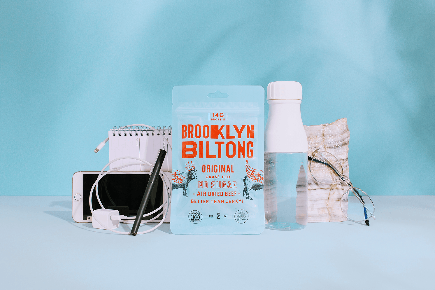 Light blue Brooklyn Biltong Original packaging with orange typography, styled with a phone, notebook, pen, water bottle, and glasses against a blue background.