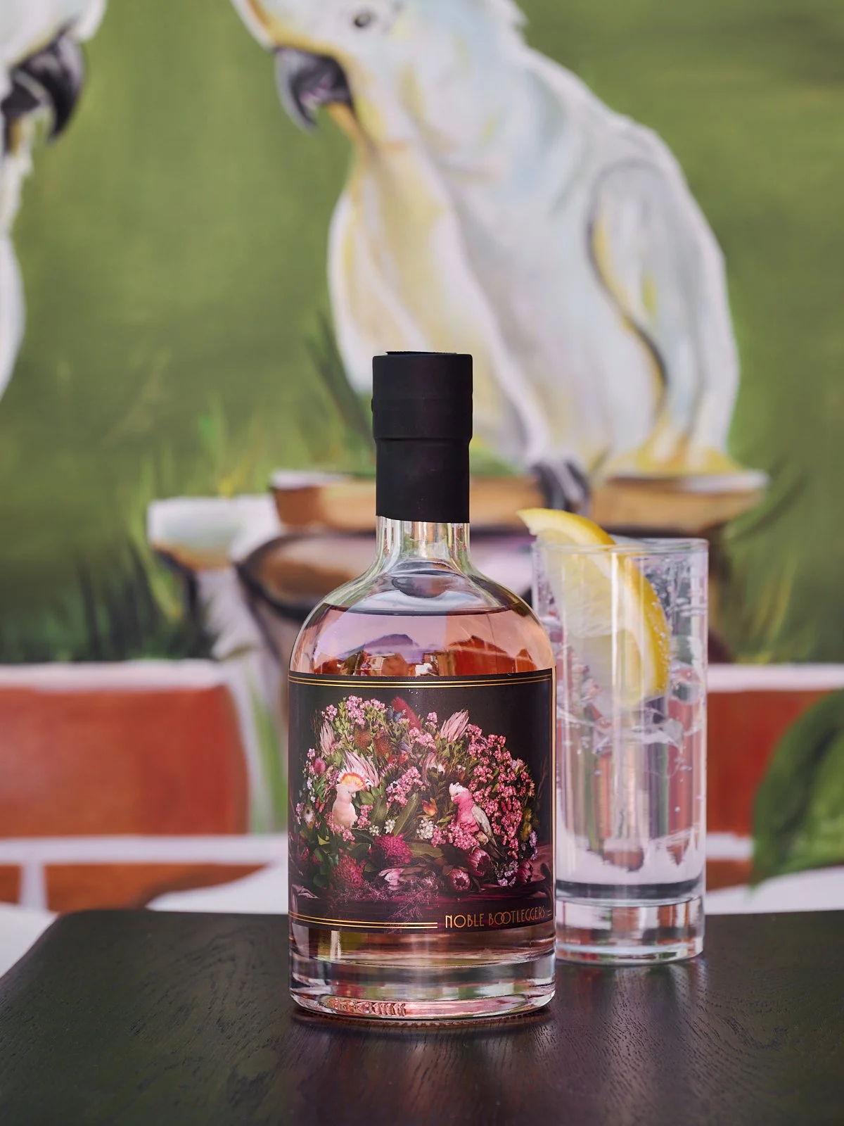 Noble Bootleggers Pink Native Gin, accompanied with a tall glass of Pink Native & tonic water, ganished with  a slice of lemon. Background contains a painted image of a cockatoo