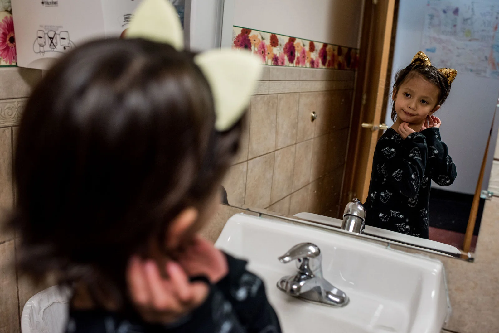 Age 6. He goes to the bathroom at a birthday party and uses the girls’ restroom. The cat ears headband is a party favor. He checks his ears in the mirror before returning to the party.