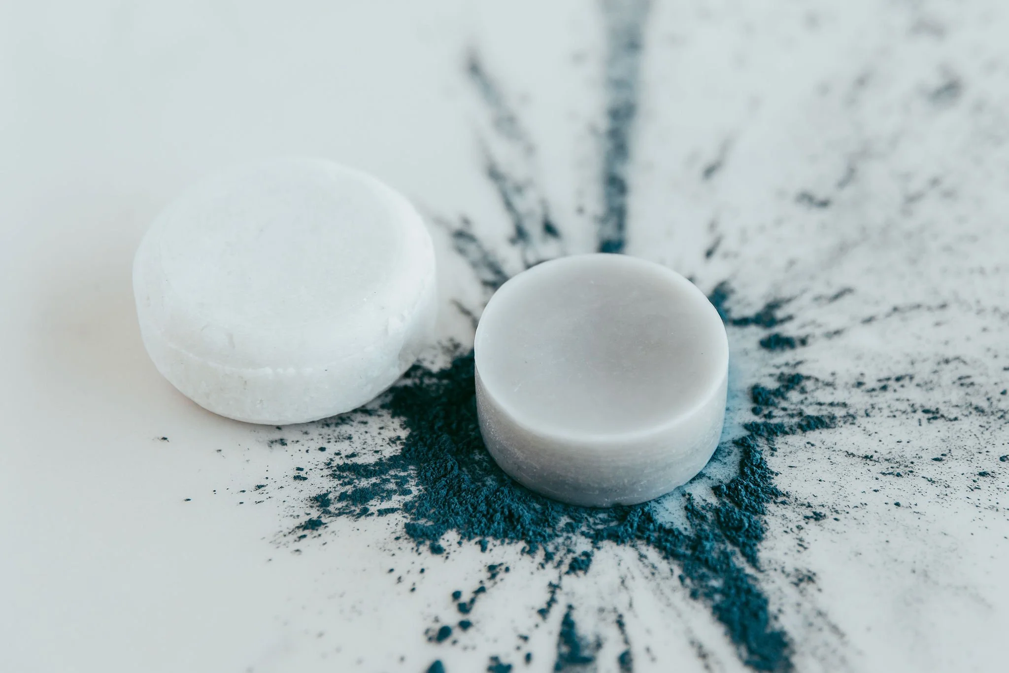 One Shampoo Bar and one conditioner bar on a white surface with natural indigo powder splattered around them.