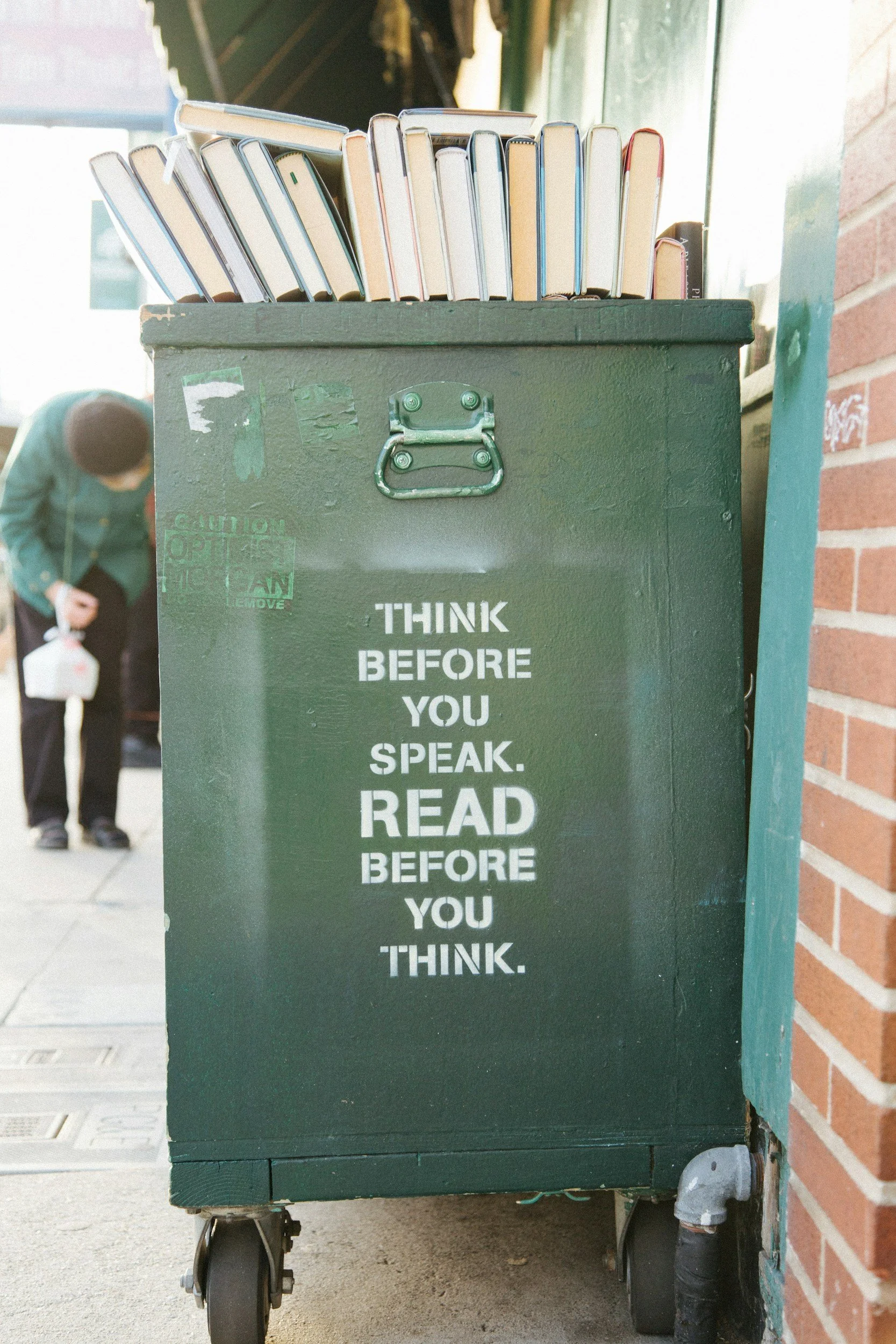 A green cart with a hinged lid and black wheels, featuring a white stencil message: "Think before you speak. Read before you think." On top of the cart, there are books standing upright.