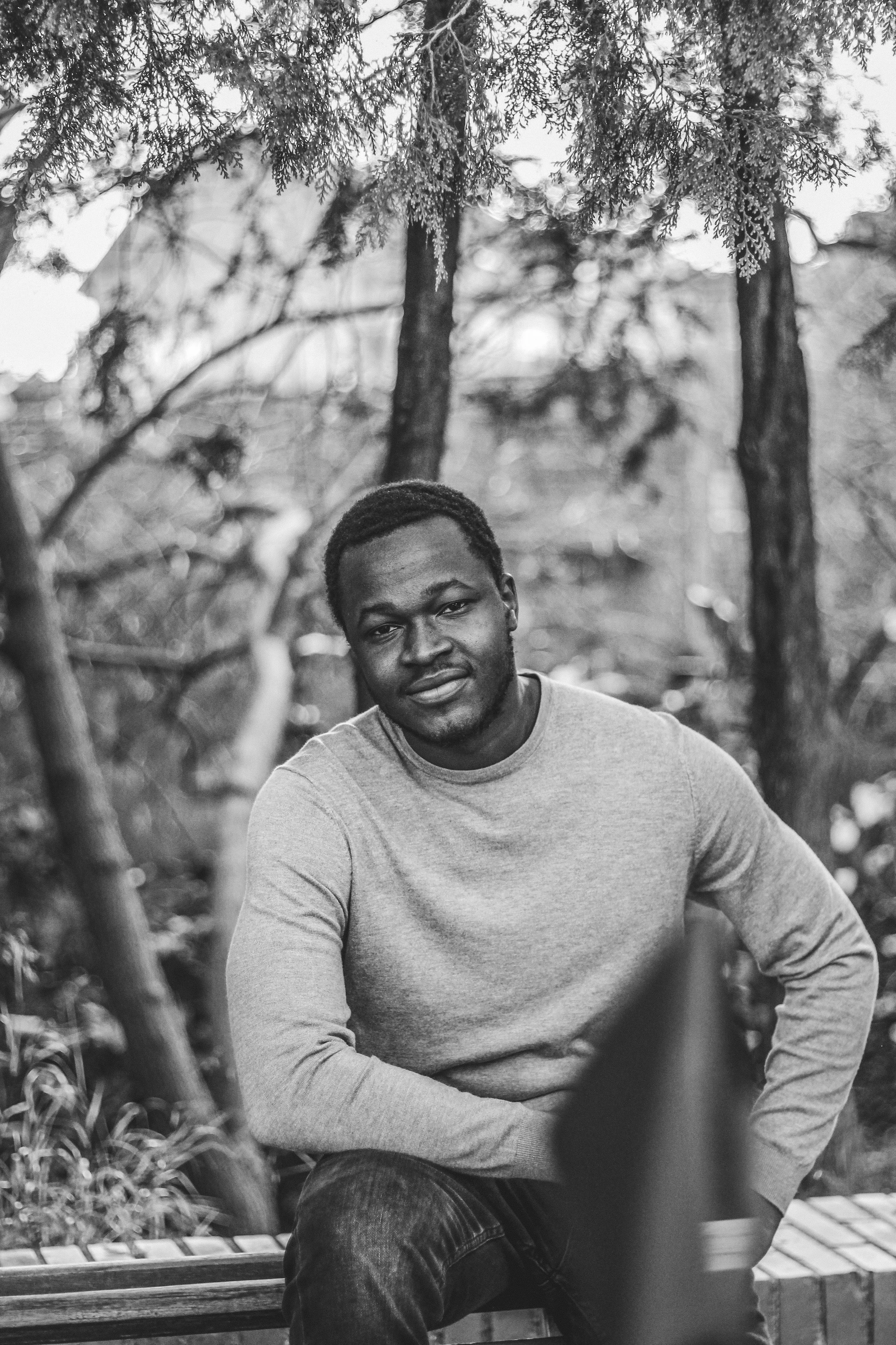 Black and white photo of a handsome, Black man in his 30s or 40s, wearing a slight smile, grey sweater and blue jeans. He is sitting on a bench, in front of a background of trees.