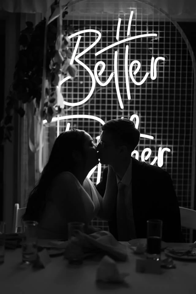Photo of a heterosexual, Latino couple in black and white silhouette. They are sitting in front of a neon "Better Together" sign and kissing. Photo by Joeyy Lee via Unsplash.com