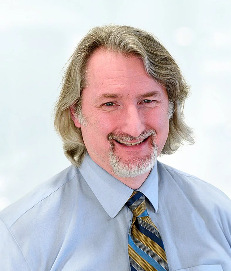 A headshot photograph of Allan Mills wearing a blue shirt and striped tie.