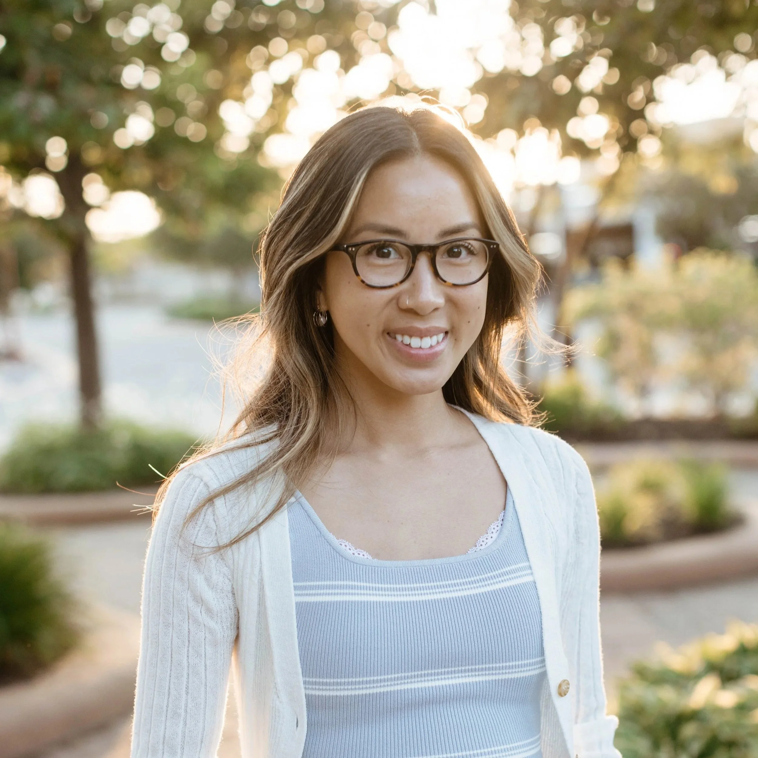 A successful experience design consultant with brown hair, wearing glasses and a white cardigan, smiling outdoors during golden hour with sunlight filtering through trees in the background.