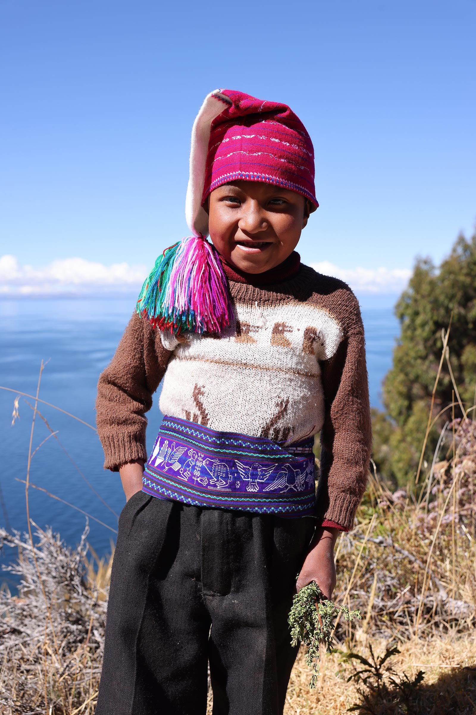En pojke i rosa mössa och traditionella kläder som precis plockat muña han har i handen, på ön Taquile i Titicacasjön, Peru