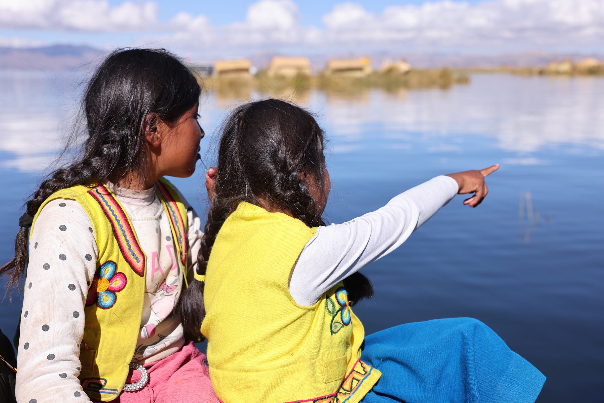 Två flickor med långa svarta flätor och gula västar från urosfolket. Den ena flickan pekar ut över Titicacasjön och Uros flytande vassöar, Peru.