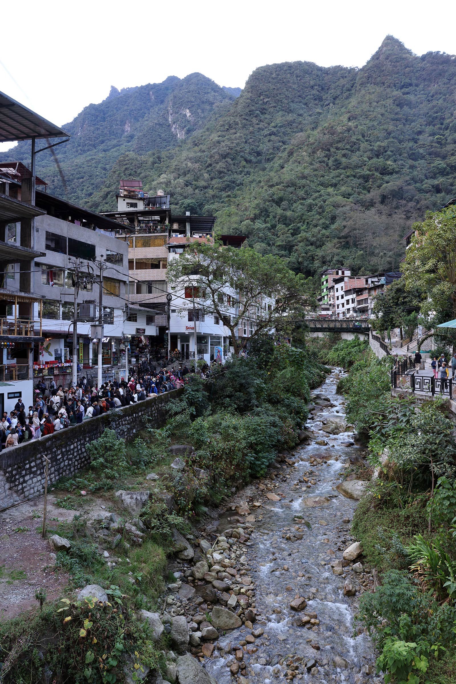 Ett vattendrag, grönskande berg och en lång kö i väntan på bussen till Machu Picchu i samhället Machu Picchu Pueblo/Aguas Calientes, Peru