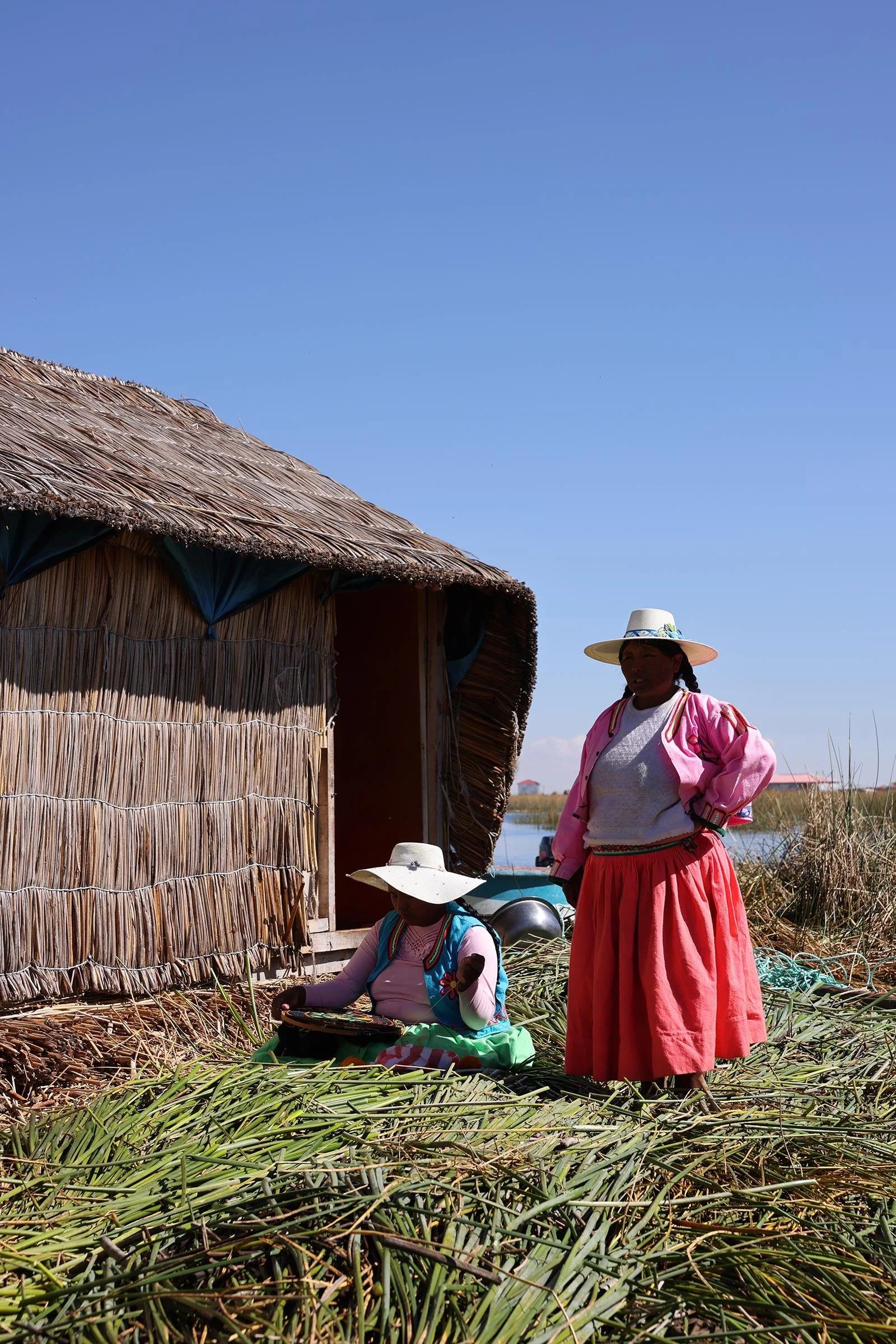 Två kvinnor, varav den ena håller på med handarbete, framför ett vasshus på en av Uros flytande vassöar i Titicacasjön, Peru