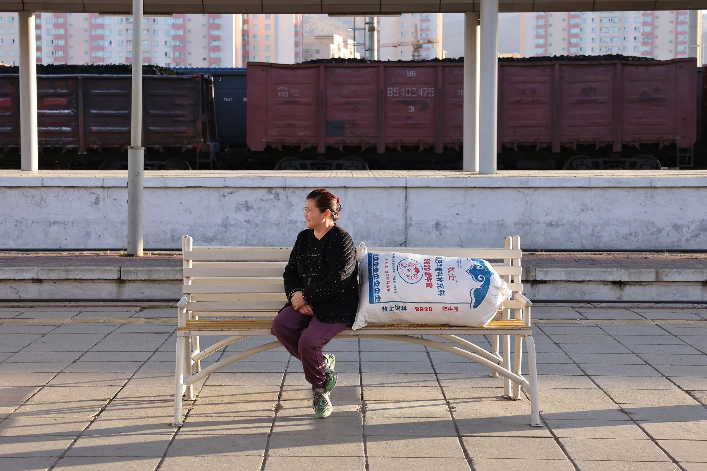 En kvinna sitter och väntar i kvällsljuset på en bänk på perrongen till Ulaanbaatars järnvägsstation. Hon har en säck djurfoder med sig och i bakgrunden syns godståg med kol. Ulaanbaatar, Mongoliet.