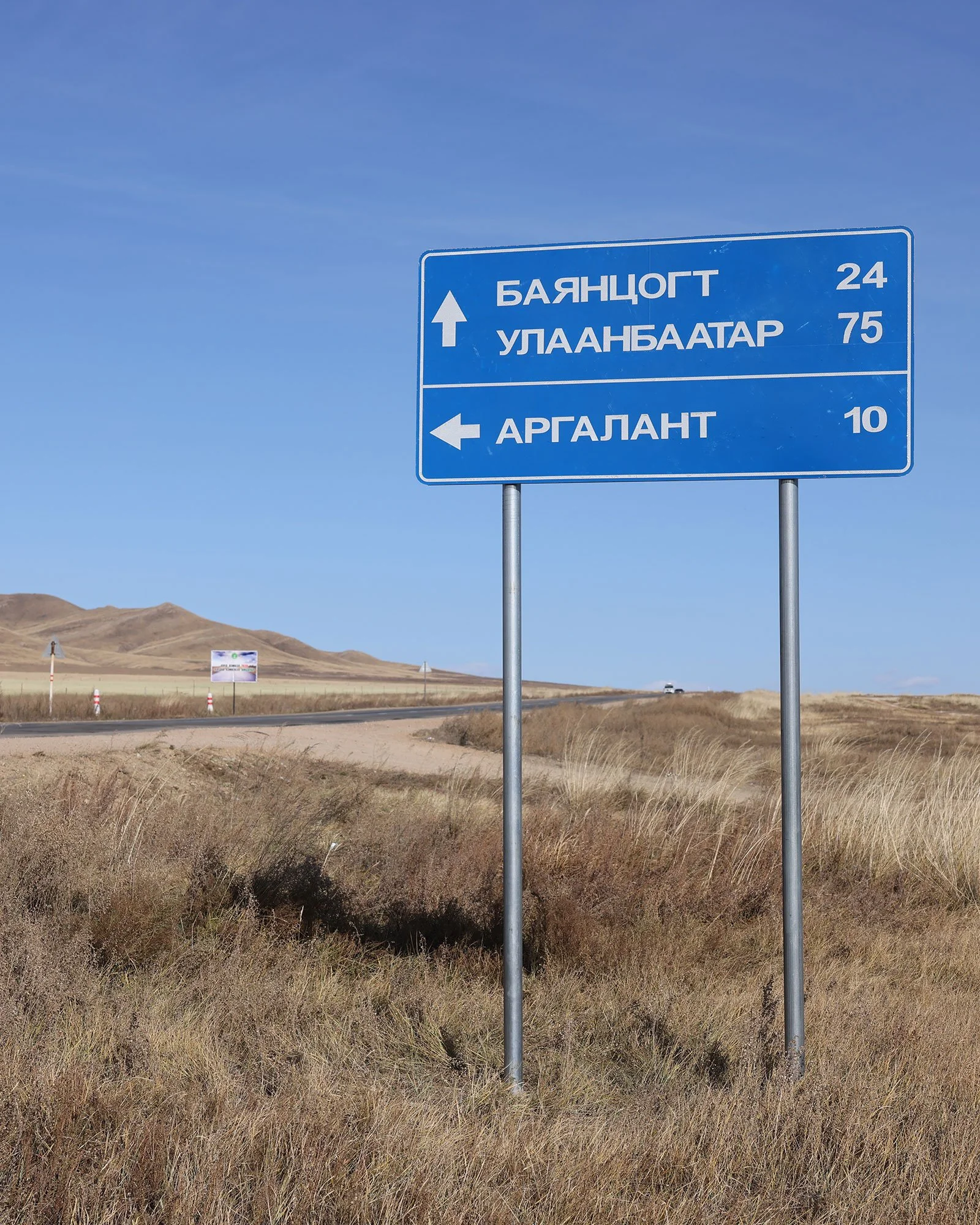Blå vägskylt i Mongoliet med kyrilliska bokstäver där det bland annat står att det är sjuttiofem kilometer till Ulaanbaatar