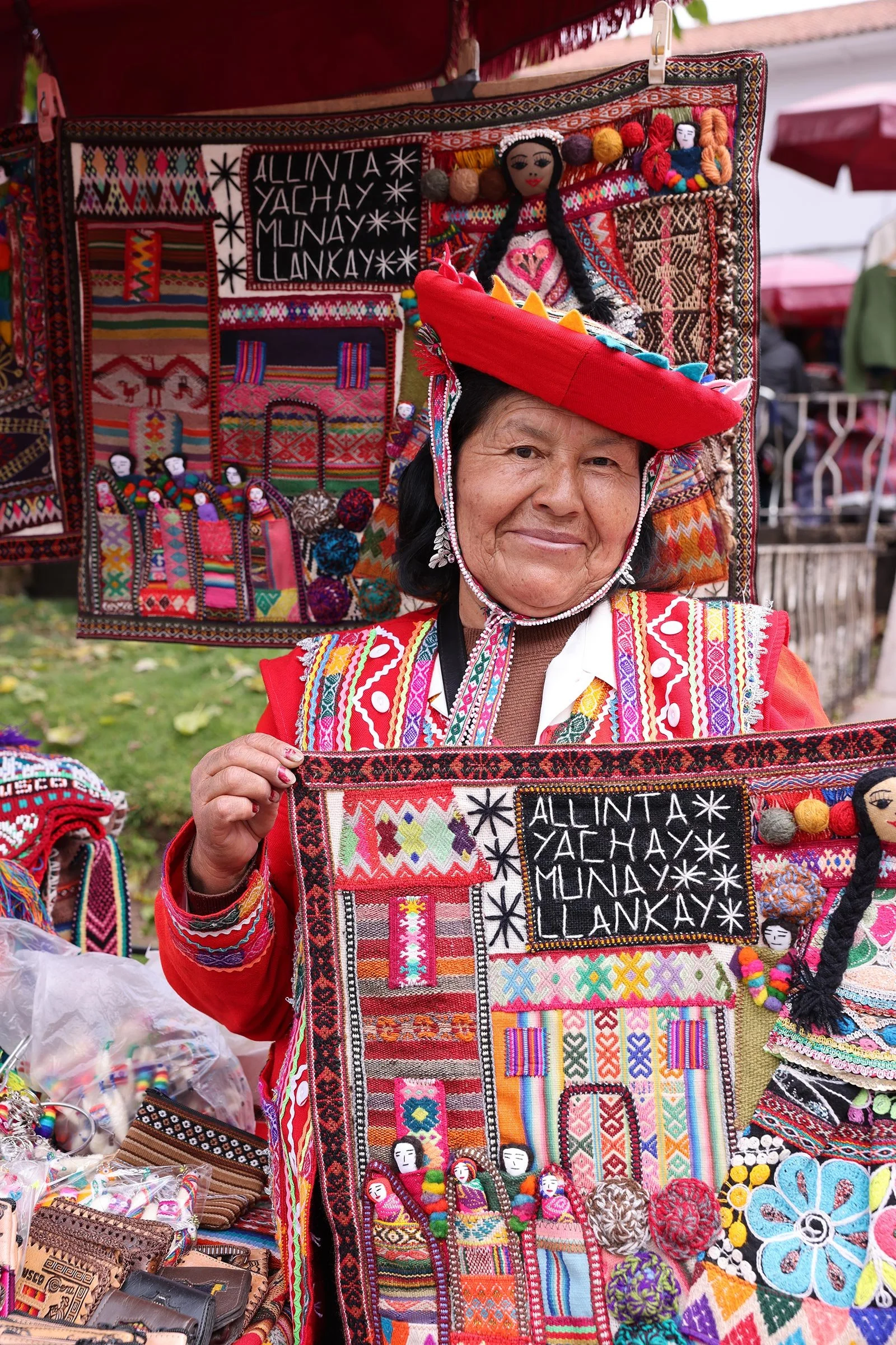 En kvinna i traditionella kläder och röd hatt håller upp peruanskt hantverk i form av en väggbonad i Cusco, Peru