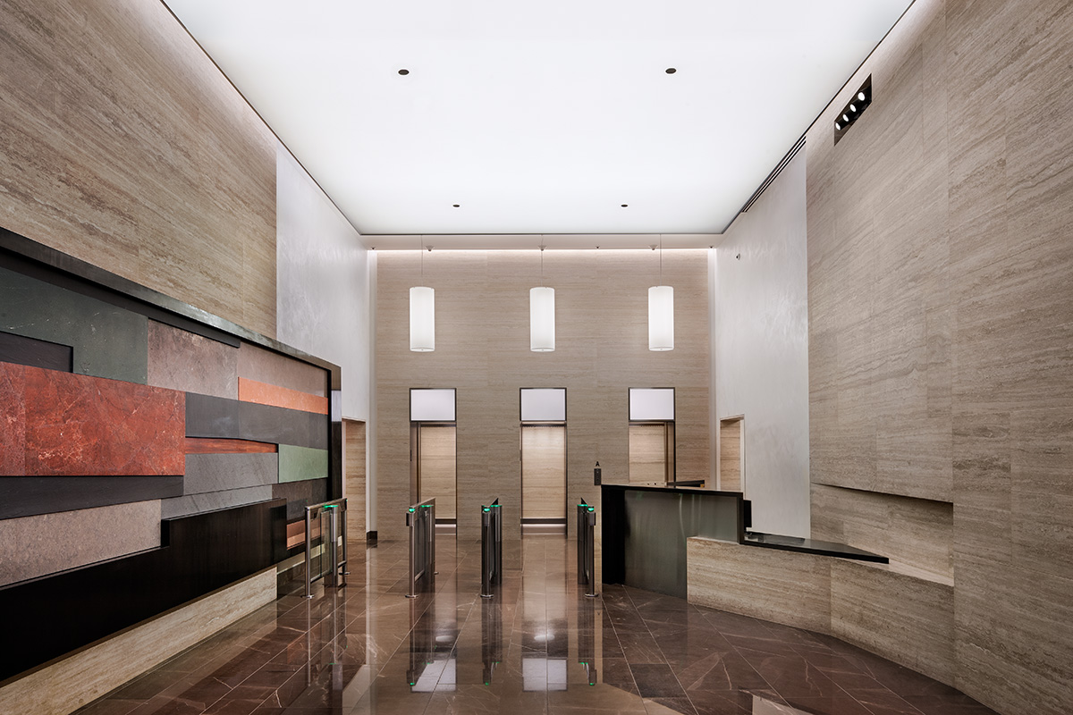 Trinity Real Estate asked us to invigorate and give a fresh identity to a very dated lobby to attract new “hip” tenants appropriate to the changing Hudson Square neighborhood. However they also wanted the design to be “timeless”: appropriate to a client with a long history in Lower Manhattan. We approached the design as an assembly of minimalist and crisply detailed wall and ceiling surfaces made of timeless materials serving as a backdrop for two dramatic sculptural elements. One is a 40’ long by 8’ high composition of different size slabs of marble, granite, limestone, slate, and metal; bordered with a giant oiled mill steel irregularly shaped finish frame, and hung off a projection on one of two side walls. The second sculptural element is a reception desk comprised of the same materials that appears to grow out of another wall projection, on the opposite wall. The walls behind each are new floor to ceiling (14’ high) horizontally grained limestone. A translucent glowing backlit stretched ceiling membrane creates an even glow throughout the lobby, and is echoed by large new call lanterns above the existing elevator doors. By selectively eliminating window mullions and putting in larger pieces of glass, we created a more open view in and out of the existing entry. Because the entrance doors are recessed from the front of the building, a new stainless steel canopy was designed as a minimalist open frame without a roof, extending out over the sidewalk. New elevator cab interiors also reflected the material palette of the lobby.