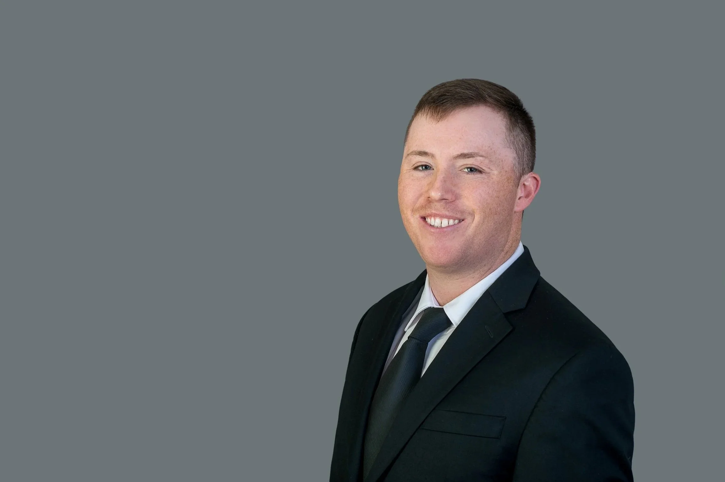Portrait of a young man with short brown hair, wearing a black suit and tie, smiling against a gray background.