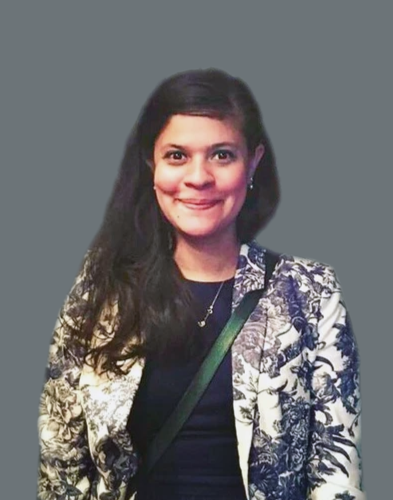 A woman with long dark hair, wearing a patterned blazer over a dark top, and smiling at the camera, against a plain gray background.