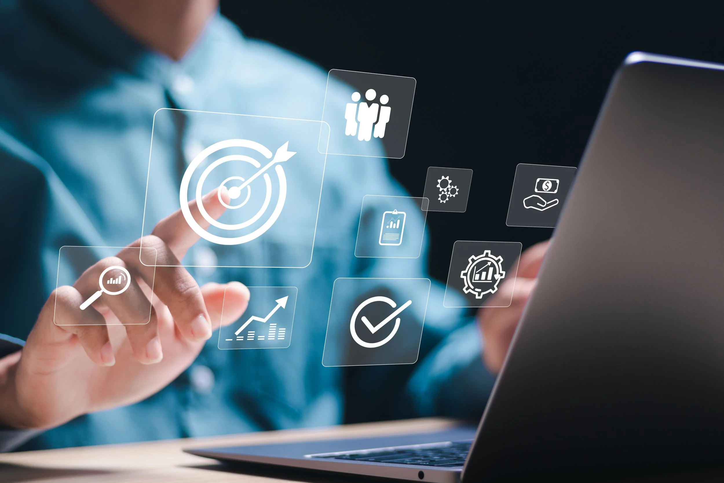 Person using a laptop with transparent digital icons related to targets, teamwork, money, and growth floating above their hand.