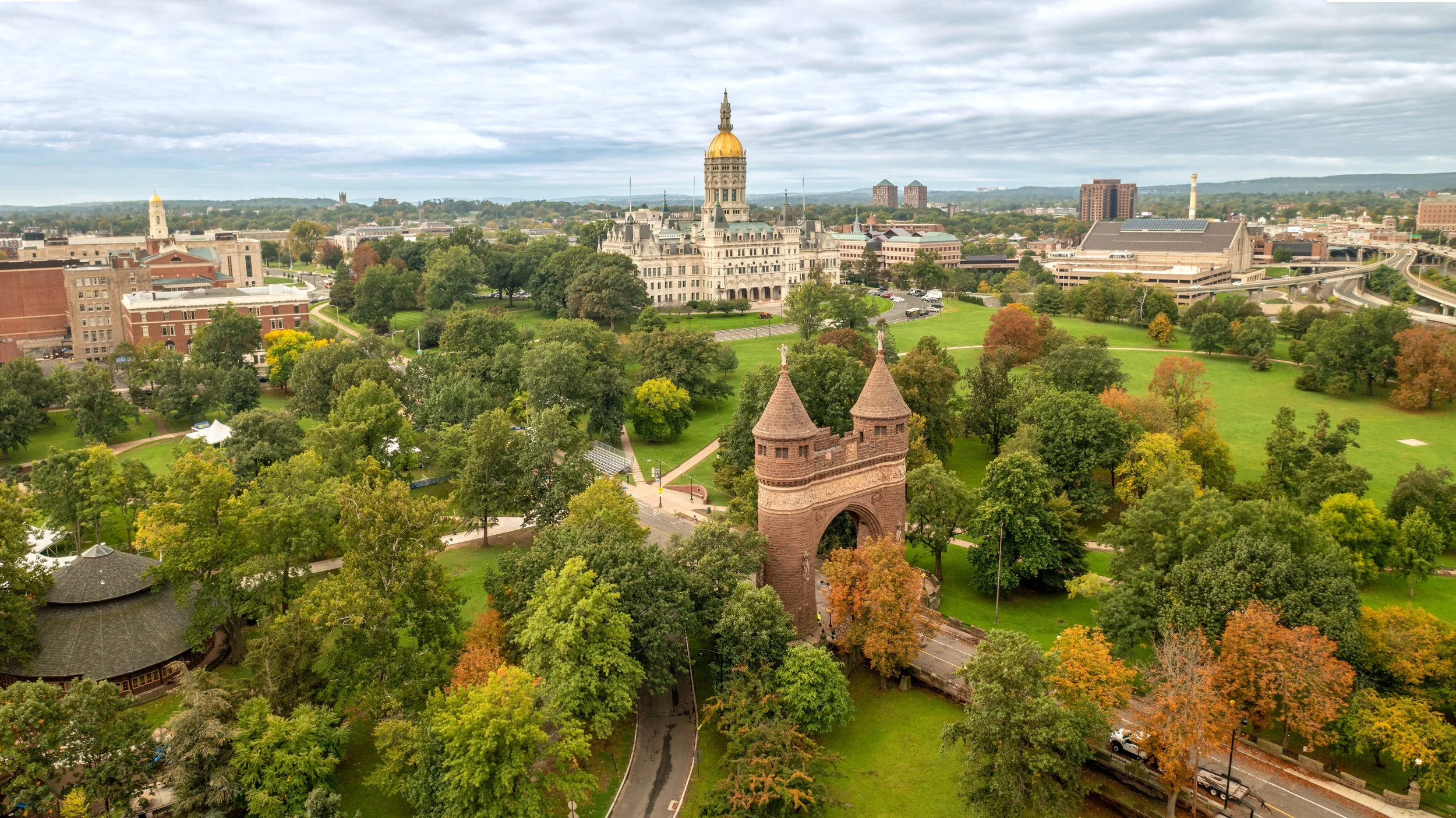 Aerial view of a Hartford, Connecticut with lush green trees and pathways, historic buildings, and a clock tower, with more city buildings and hills in the background under a cloudy sky.