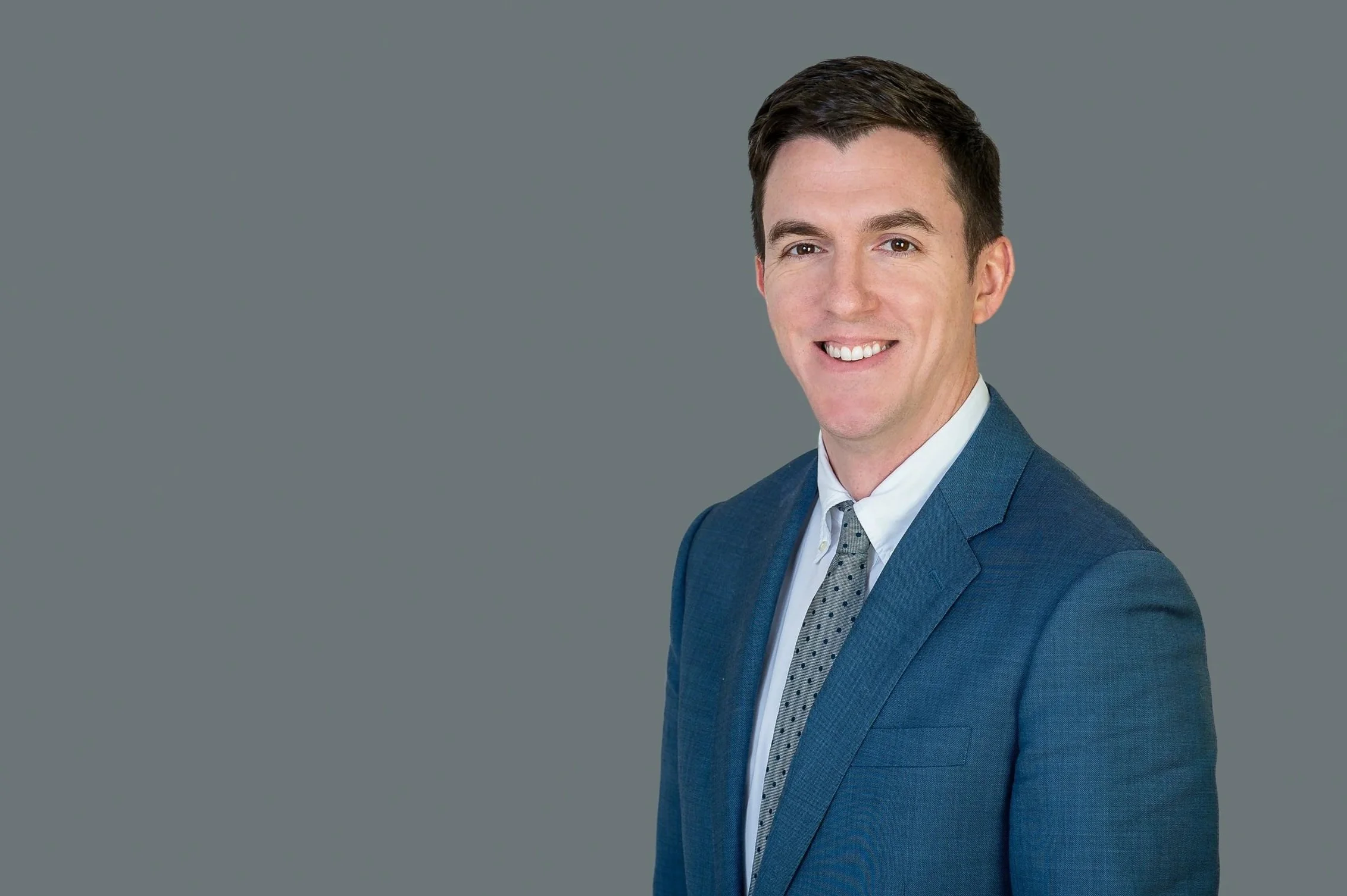Portrait of John Aliberti with dark hair and a bright smile, wearing a blue suit, white shirt, and patterned tie, against a gray background.